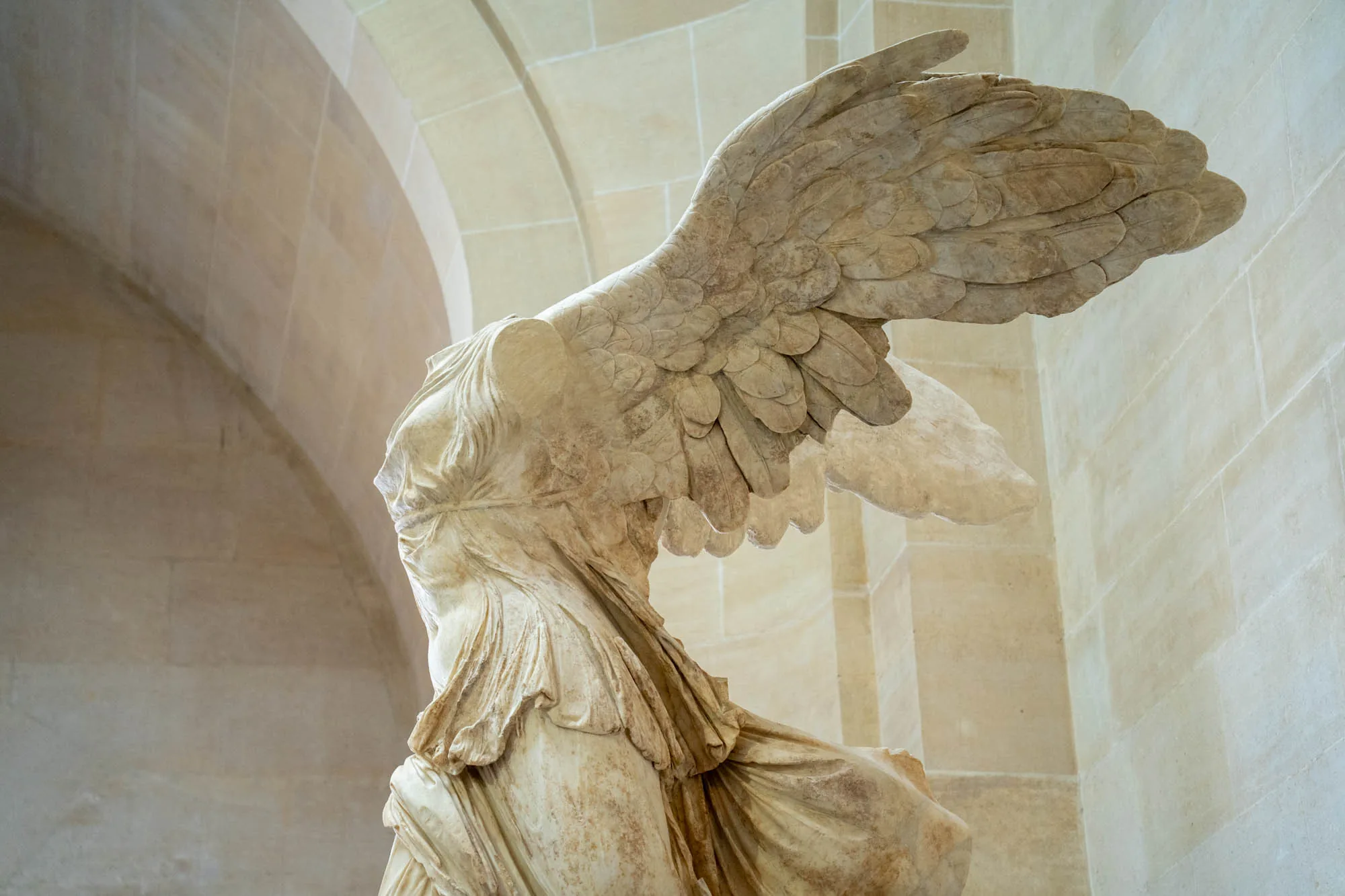 写真はフランスのパリにあるルーブル美術館に展示されている、有名な彫刻「サモトラケのニケ」を捉えています。大理石でできた像は、翼を広げた勝利の女神ニケを表現しており、その躍動感と繊細な造形が印象的です。像は頭部と両腕を欠損していますが、その荘厳な佇まいは見る者を圧倒します。背景にはベージュ色の石造りの壁とアーチ状の天井が見え、美術館の落ち着いた雰囲気を醸し出しています。光の当たり具合が、像の質感や陰影を際立たせ、より立体的に見せています。