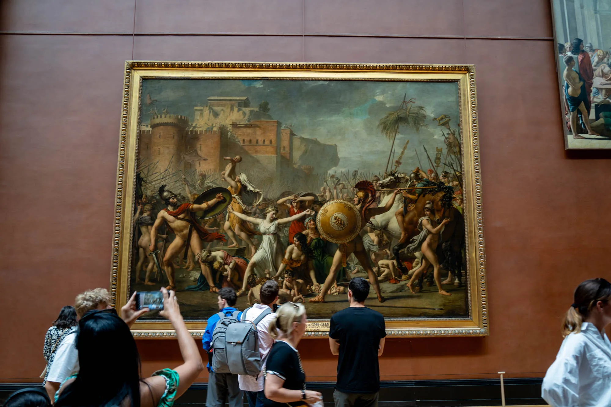写真はフランスのパリにあるルーブル美術館で撮影されたものと思われます。画面の中央には、金色の豪華な額縁に飾られた大きな絵画が飾られています。絵画は、古代の都市を背景に、裸体またはそれに近い姿の人物たちが争っている様子を描写しています。画面手前には、数人の鑑賞者が絵画を鑑賞しており、スマートフォンで写真を撮影している人もいます。絵画の雰囲気は古代の戦いをテーマにしており、背景の建築物や人物の衣装から古代ギリシャまたはローマを連想させます。絵画の色調はやや暗めで、鑑賞者のいる空間は明るく、絵画鑑賞に集中できるようになっています。