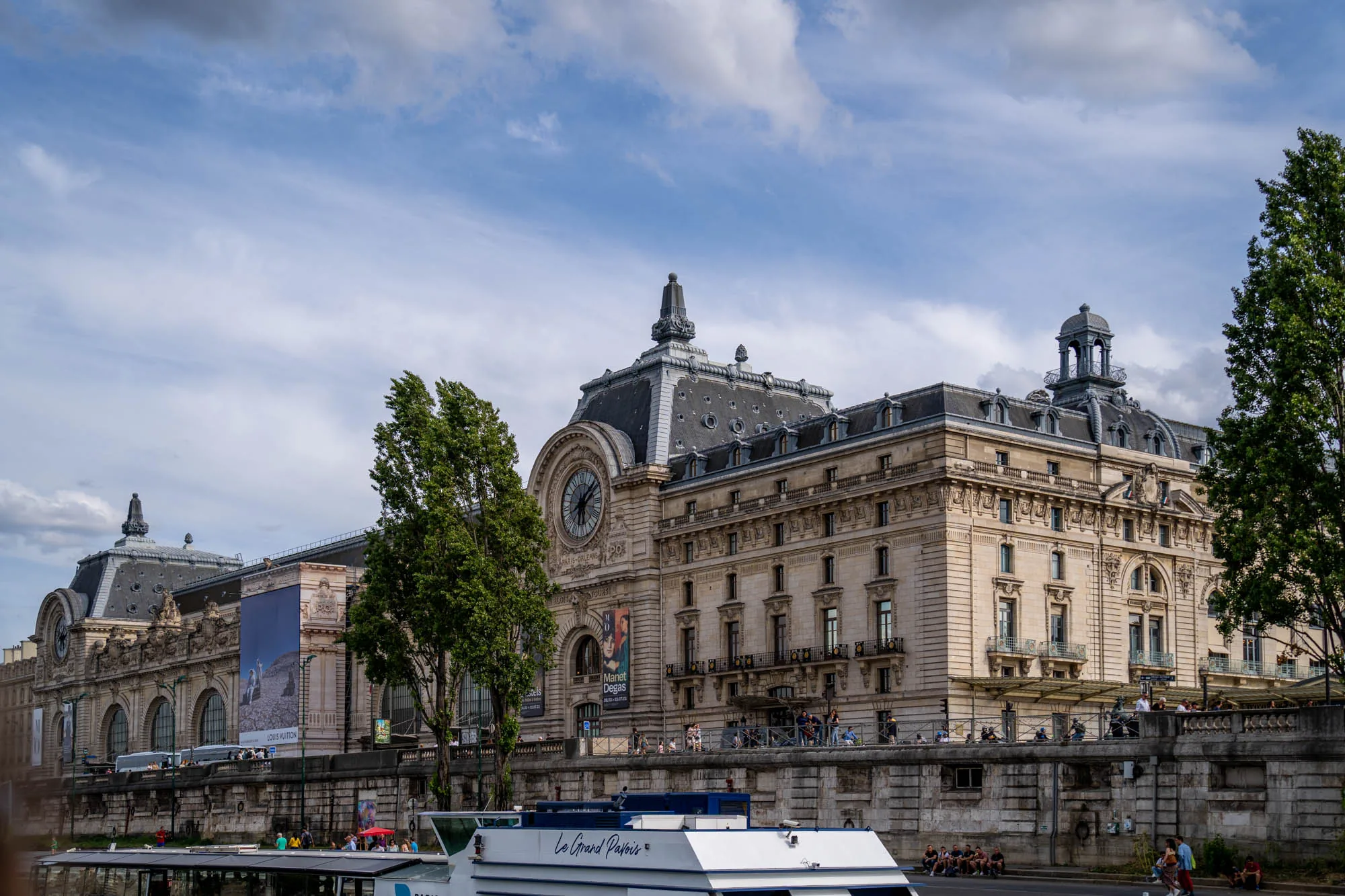 写真には、フランスのパリにあるオルセー美術館が写っています。セーヌ川から撮影されたと思われる画像には、美術館の美しい建物が大きく写っています。石造りの外観は壮大で、時計台や装飾的な屋根のディテールが印象的です。建物の壁には、展示会のポスターやルイ・ヴィトンの広告が掲示されています。美術館前の遊歩道には、多くの人々がおり、景色を眺めたり、くつろいだりしている様子が伺えます。手前にはセーヌ川を航行する観光船の一部が写っており、"Le Grand Pavois"という船名が確認できます。空は晴れており、いくつかの雲が浮かんでいます。全体的に、パリの美しい風景とオルセー美術館の文化的魅力が伝わってくる写真です。
