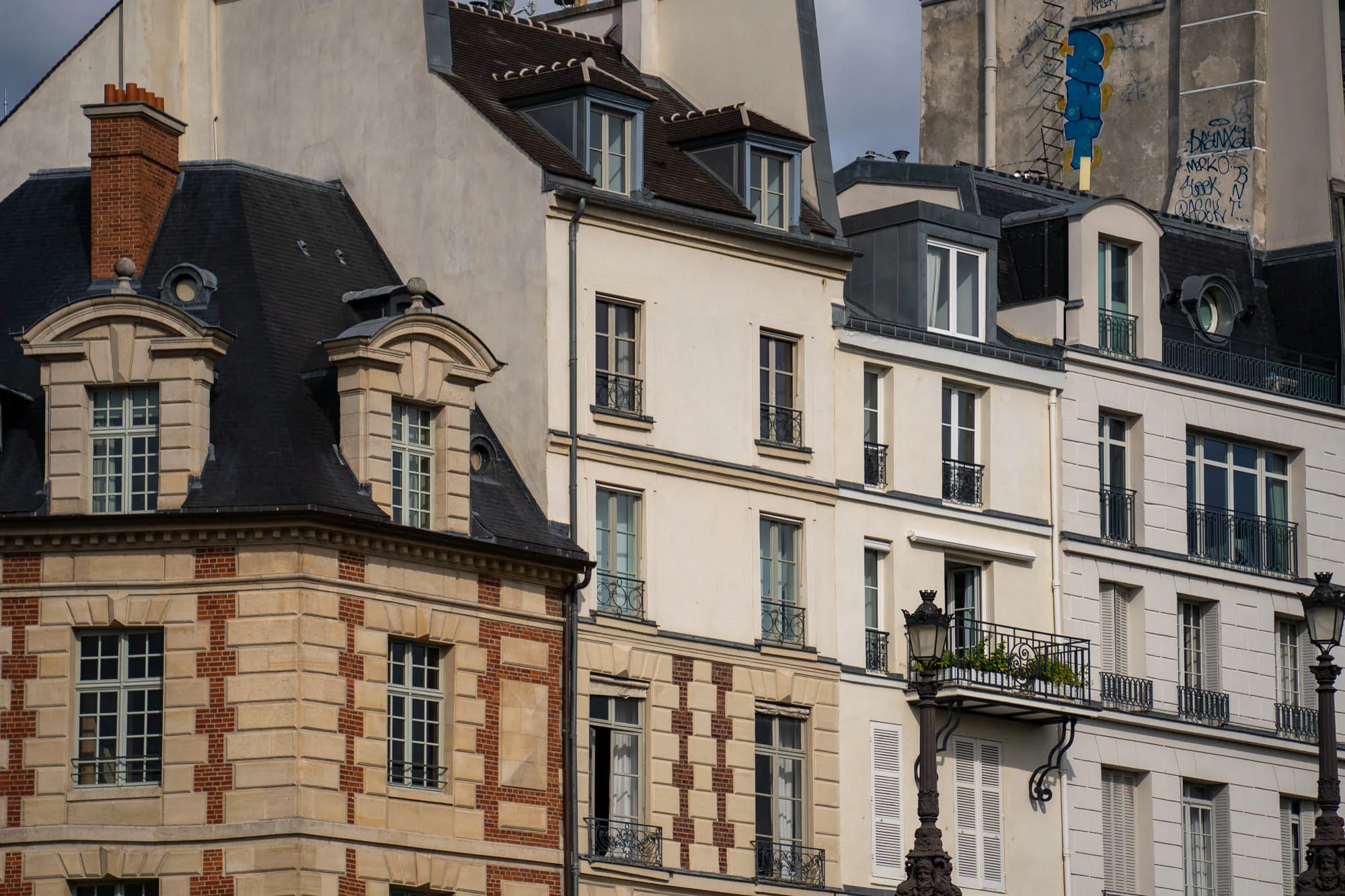 写真には、フランス・パリの街並みが写っています。複数の建物が隣接しており、それぞれ異なる建築様式を見せています。左端の建物は、ベージュ色の石材と赤いレンガの装飾が組み合わされており、クラシックな雰囲気を醸し出しています。屋根は黒色で、窓枠も整然と配置されています。中央の建物は、クリーム色の外壁で、ややシンプルなデザインです。窓には小さなバルコニーが付いているものもあります。右側の建物は、よりモダンな印象で、バルコニーには緑の植物が飾られています。最上階には落書きが見られます。全体的に、パリらしい洗練された美しさと、歴史を感じさせる趣が組み合わさった景観です。落ち着いた色調で、パリの街並みの静かな日常を切り取ったような雰囲気です。