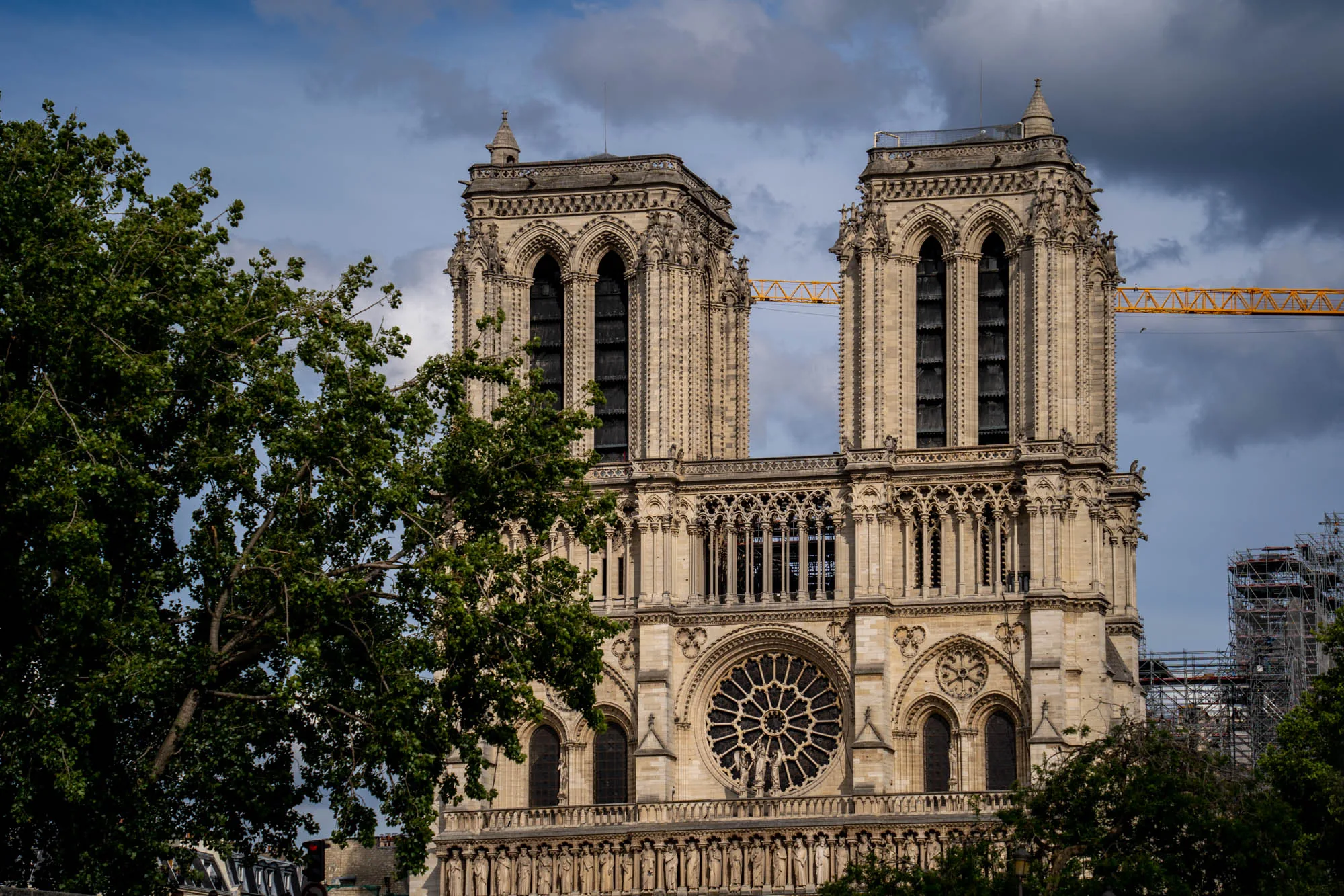 この写真はフランスのパリにあるノートルダム大聖堂を写したものです。大聖堂の正面が中心に配置され、左右には緑豊かな木々がフレームを飾っています。建物の石造りの外壁は精巧な装飾が施されており、ゴシック様式の建築様式が際立っています。特に印象的なのは中央にある大きなバラ窓で、複雑なデザインが目を引きます。写真全体を通して、空には雲が広がり、少し陰影のある雰囲気を出しています。背景には工事用のクレーンや足場が見え、大聖堂が修復中であることがわかります。この写真は、歴史的な建造物の荘厳さと、復興への道のりを捉えた一枚と言えるでしょう。