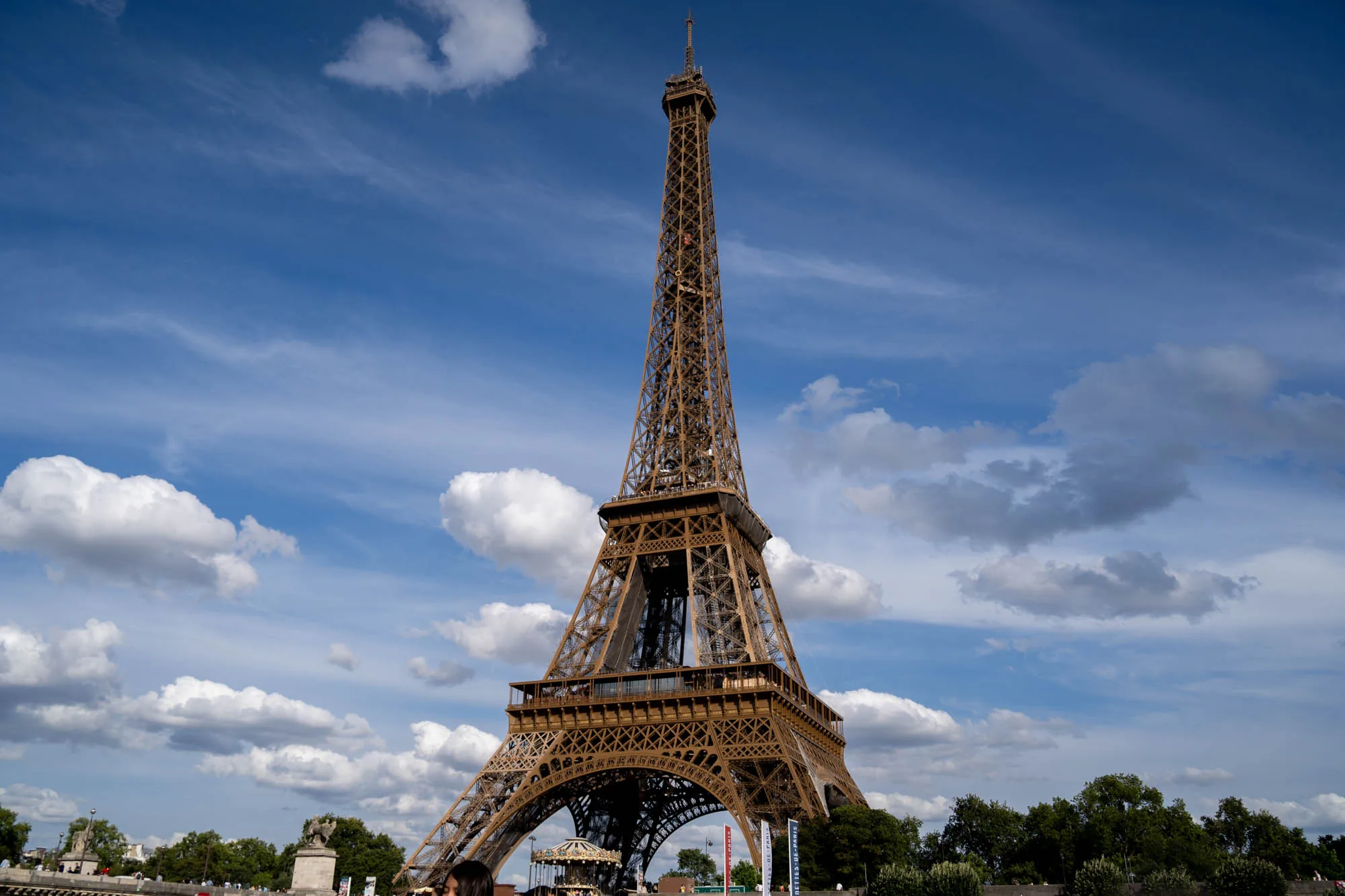 フランスのパリで撮影された写真です。青空を背景に、堂々とそびえ立つエッフェル塔が大きく写っています。空には白い雲が浮かび、塔の複雑な鉄骨構造を際立たせています。エッフェル塔の足元には木々やメリーゴーランドの一部が見え、観光地としての賑わいを伝えています。全体的に明るく爽やかな雰囲気で、パリの象徴的な風景を捉えた一枚です。