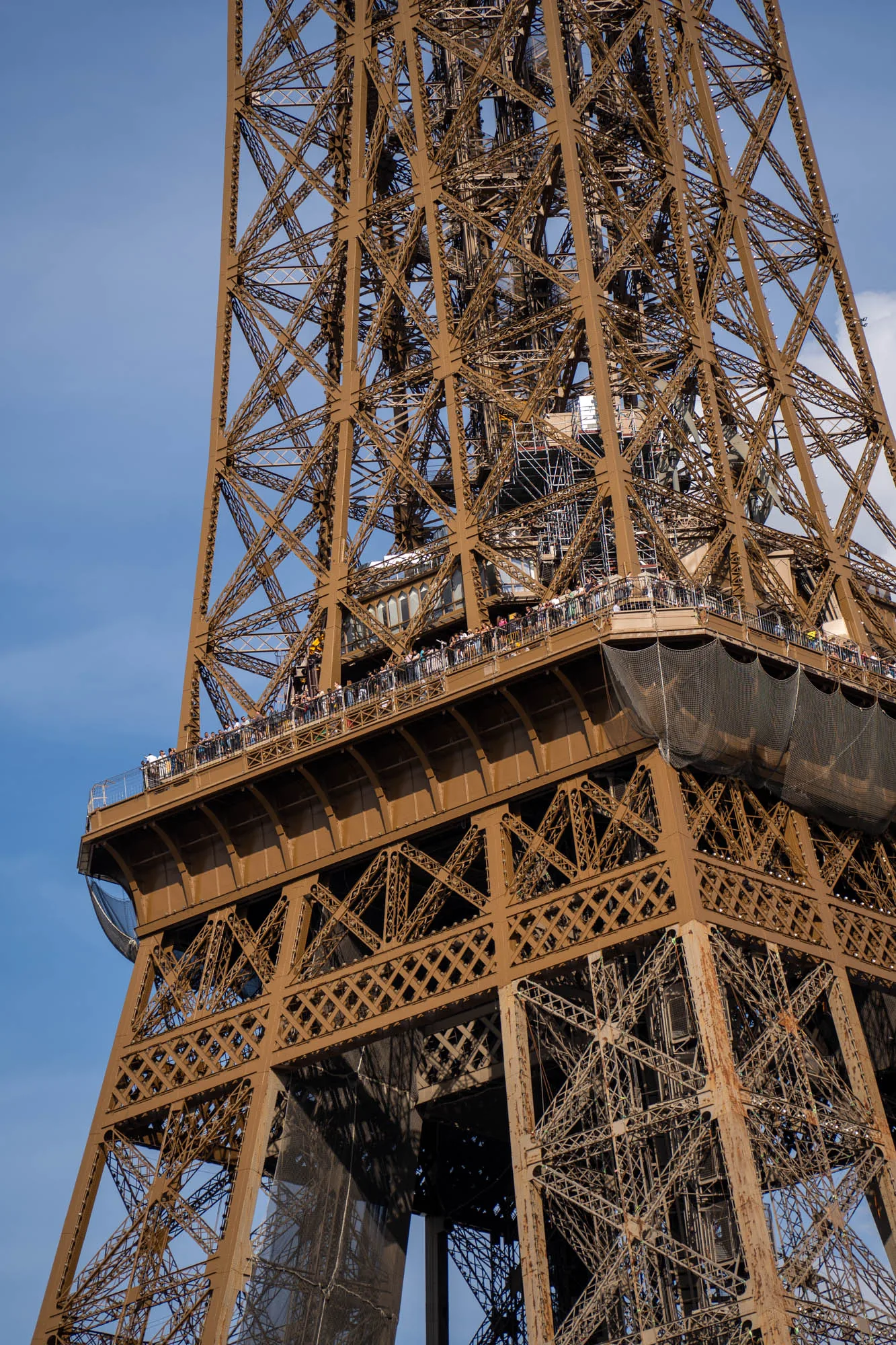 フランスのパリで撮影された写真です。画面いっぱいにエッフェル塔の下から見上げた部分が写っています。エッフェル塔の鉄骨が複雑に組み合わさっており、その構造美が際立っています。一部には展望台があり、多くの観光客が景色を眺めている様子が見えます。鉄骨の間からは青空が覗き、観光客がいる展望台には落下防止のネットが設置されているのが分かります。一部工事中の箇所も見られます。