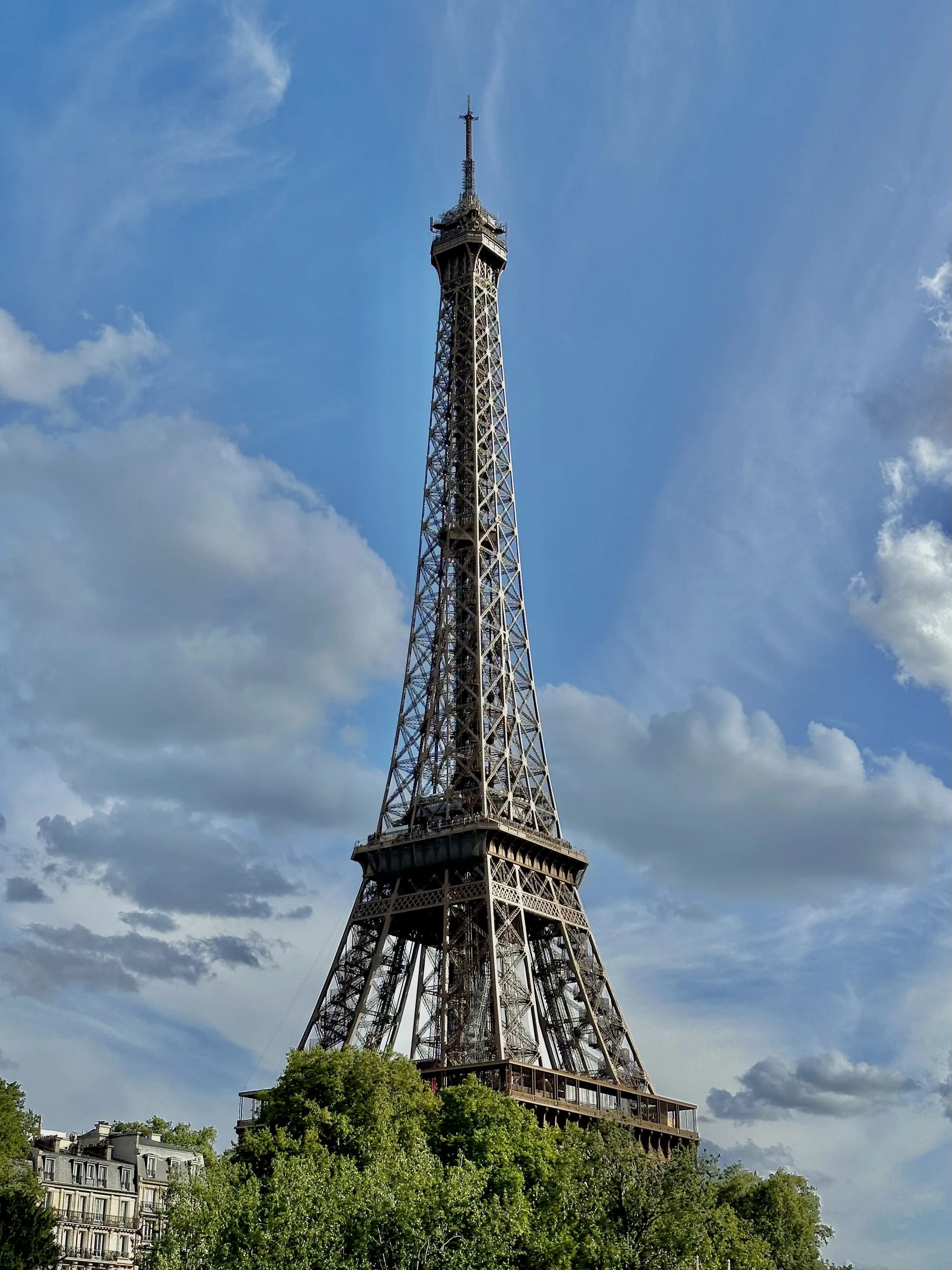 写真はフランスのパリで撮影されたエッフェル塔の全景を捉えています。エッフェル塔は画面中央に位置し、青い空と白い雲を背景にそびえ立っています。塔の鉄骨構造が緻密に表現されており、その壮大さと繊細さが伝わってきます。前景には緑豊かな樹木があり、塔の足元を覆うように茂っています。遠景にはパリの典型的な建物の一部が見え、写真に奥行きを与えています。空は晴れており、いくつかの雲が浮かんでいるため、穏やかな日であることがわかります。全体として、この写真はパリの象徴的なランドマークであるエッフェル塔の美しさと魅力を捉えたものです。