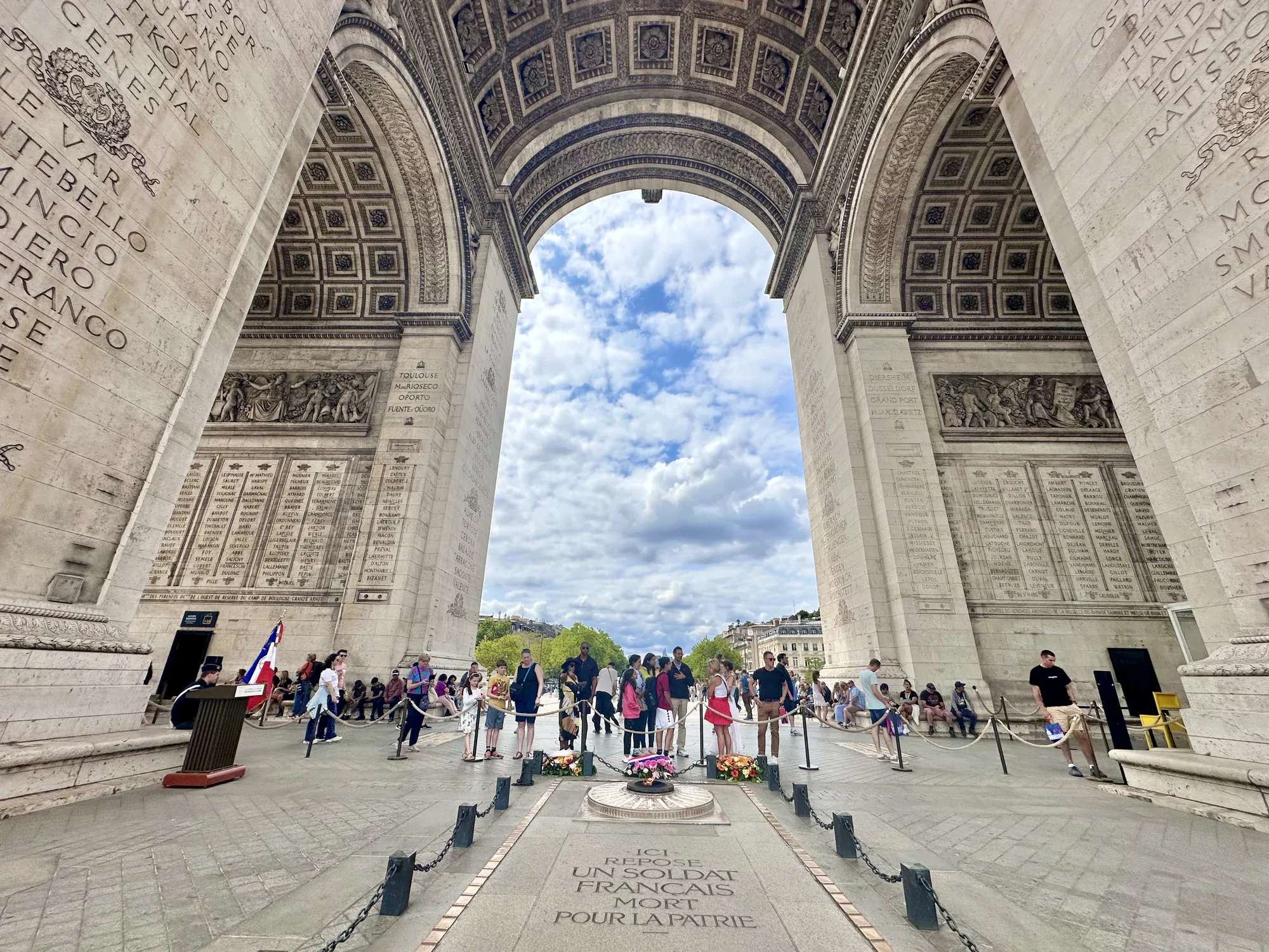 この写真はフランスのパリにある凱旋門の内部から撮影されたものです。凱旋門の壮大なアーチを通して、曇り空が見えます。凱旋門の壁には、歴史的な名前や彫刻が刻まれています。  写真の中心には、無名戦士の墓があり、その上には永遠の炎が灯っています。墓の周りには花輪が供えられており、観光客や市民がその周りを囲み、敬意を払っています。地面には「ICI REPOSE UN SOLDAT FRANÇAIS MORT POUR LA PATRIE（ここに祖国のために死んだフランス兵士が眠る）」という碑文が刻まれています。フランスの国旗を持つ警備兵が立っているのが見えます。  全体的に、この写真はパリの象徴的なランドマークである凱旋門の威厳と、無名戦士の墓に対する敬意と記憶を捉えたものです。