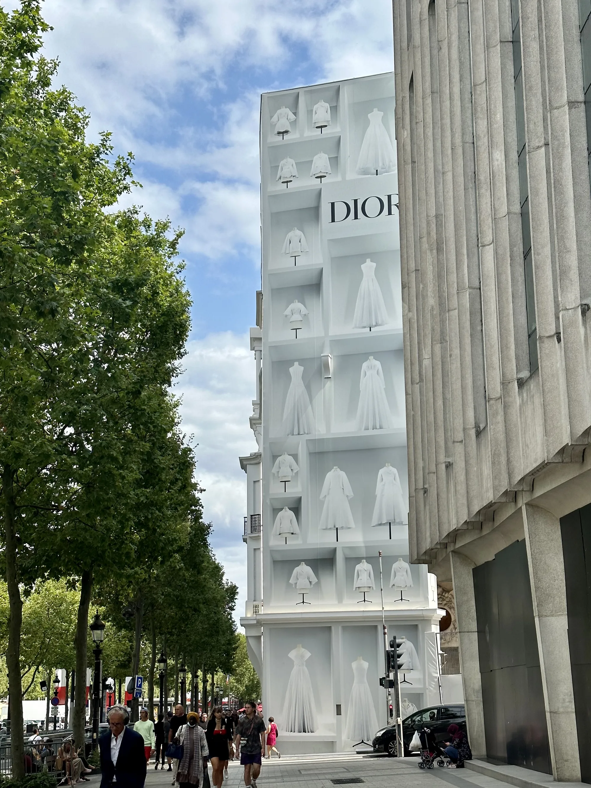 フランス、パリで撮影された写真です。青い空に白い雲が浮かび、晴れた日の街並みが写っています。左側には緑豊かな街路樹が並び、その下を人々が歩いています。中央には、白い壁にDiorのロゴと、白い衣装が飾られた建物がそびえ立っています。建物の壁面は、まるでショーケースのようにデザインされており、様々な白いドレスやジャケットがマネキンに着せられて展示されています。右側には、縦のラインが強調された重厚な印象の石造りの建物が一部写り込んでおり、歴史的な雰囲気を感じさせます。通りには車が走っており、人々の日常の風景が垣間見えます。全体的に、洗練されたファッションとパリの美しい街並みが調和した、エレガントな雰囲気を醸し出しています。