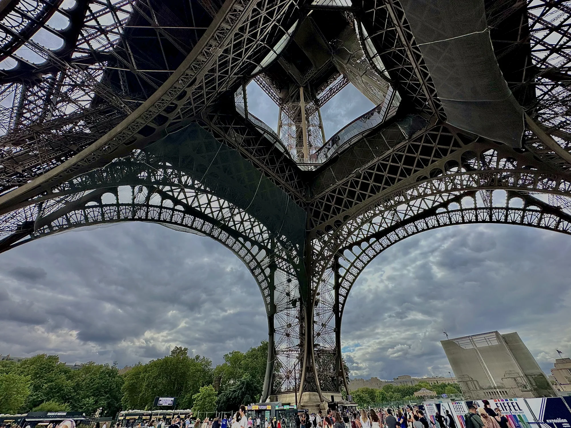 この写真はフランスのパリで撮影されたもので、エッフェル塔の真下から空を見上げているような構図です。画面全体を覆うようにそびえ立つエッフェル塔の鉄骨が、緻密な幾何学模様を描いています。鉄骨の間から見える空は、厚い雲に覆われており、やや暗い印象を与えます。  写真の下部には、エッフェル塔の足元に集まる大勢の人々が写っています。様々な服装をした人々が、思い思いに観光を楽しんでいる様子が伺えます。背景には、緑豊かな木々や他の建造物が見え、パリの街並みを垣間見ることができます。曇り空と相まって、写真全体に落ち着いた雰囲気が漂っています。