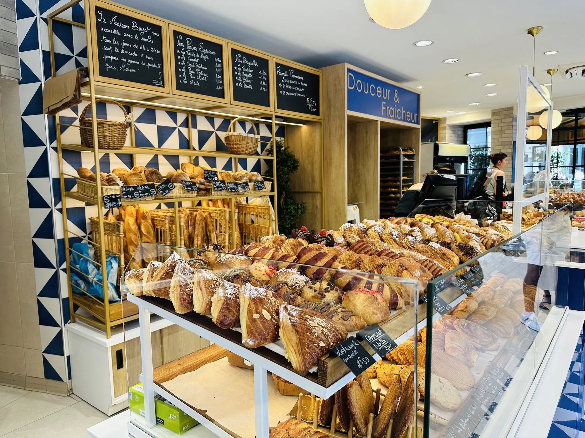 フランスのパリで撮影されたパン屋さんの写真です。店内には様々な種類のパンや焼き菓子が並べられています。手前にはクロワッサンやデニッシュなどのペストリー類がガラスケースに陳列され、奥にはバゲットやパンが棚に並んでいます。棚の背景には、青と白の幾何学模様の壁があり、おしゃれな雰囲気です。黒板にはフランス語でパンの種類と値段が書かれています。天井には丸い照明が設置されており、店内を明るく照らしています。「Douceur & Fraicheur」と書かれた看板があり、甘さと新鮮さを売りにしているようです。店員らしき女性が奥の方に見え、パンを選んでいるお客さんの姿も見えます。全体的に清潔感があり、美味しそうなパンが並んでいる様子が伝わってくる写真です。