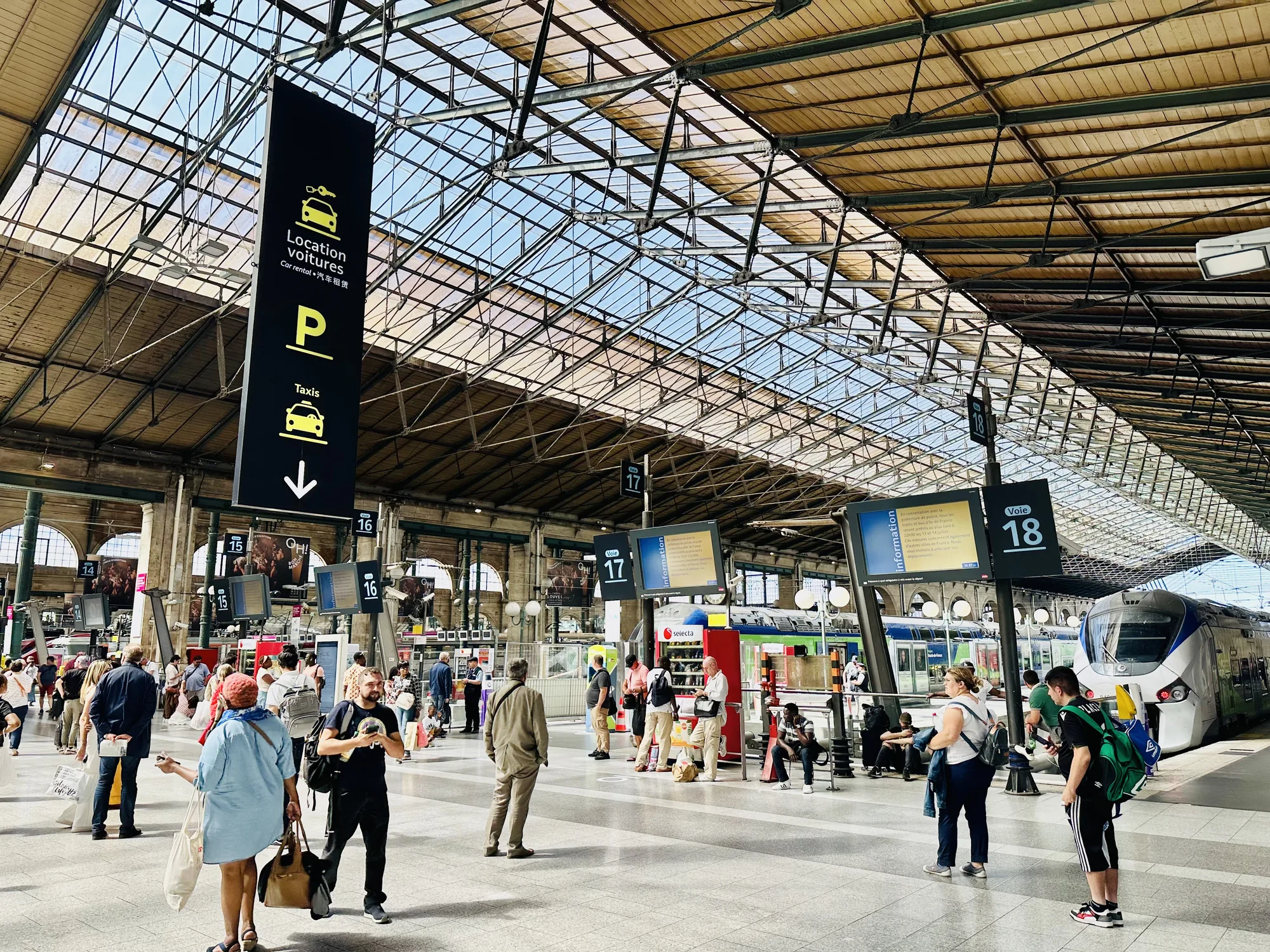 この写真はフランスのパリにある駅の構内を写したものです。天井はガラス張りで、自然光が差し込んでいます。鉄骨の構造が美しく、歴史を感じさせる雰囲気です。  構内には多くの人が行き交い、それぞれが旅行者や通勤客のように見えます。柱にはプラットフォームの番号を示す看板が掲げられており、16番、17番、18番のプラットフォームが見えます。  また、「Location voitures (レンタカー)」や「Taxis」を示す看板も吊り下げられています。出発案内板には情報が表示されており、列車の出発を待つ人々がいます。近代的な列車が停車しており、これからどこかへ向かう人々の期待感が伝わってくるようです。全体的に活気があり、旅行の始まりや終わりの瞬間を捉えた写真です。