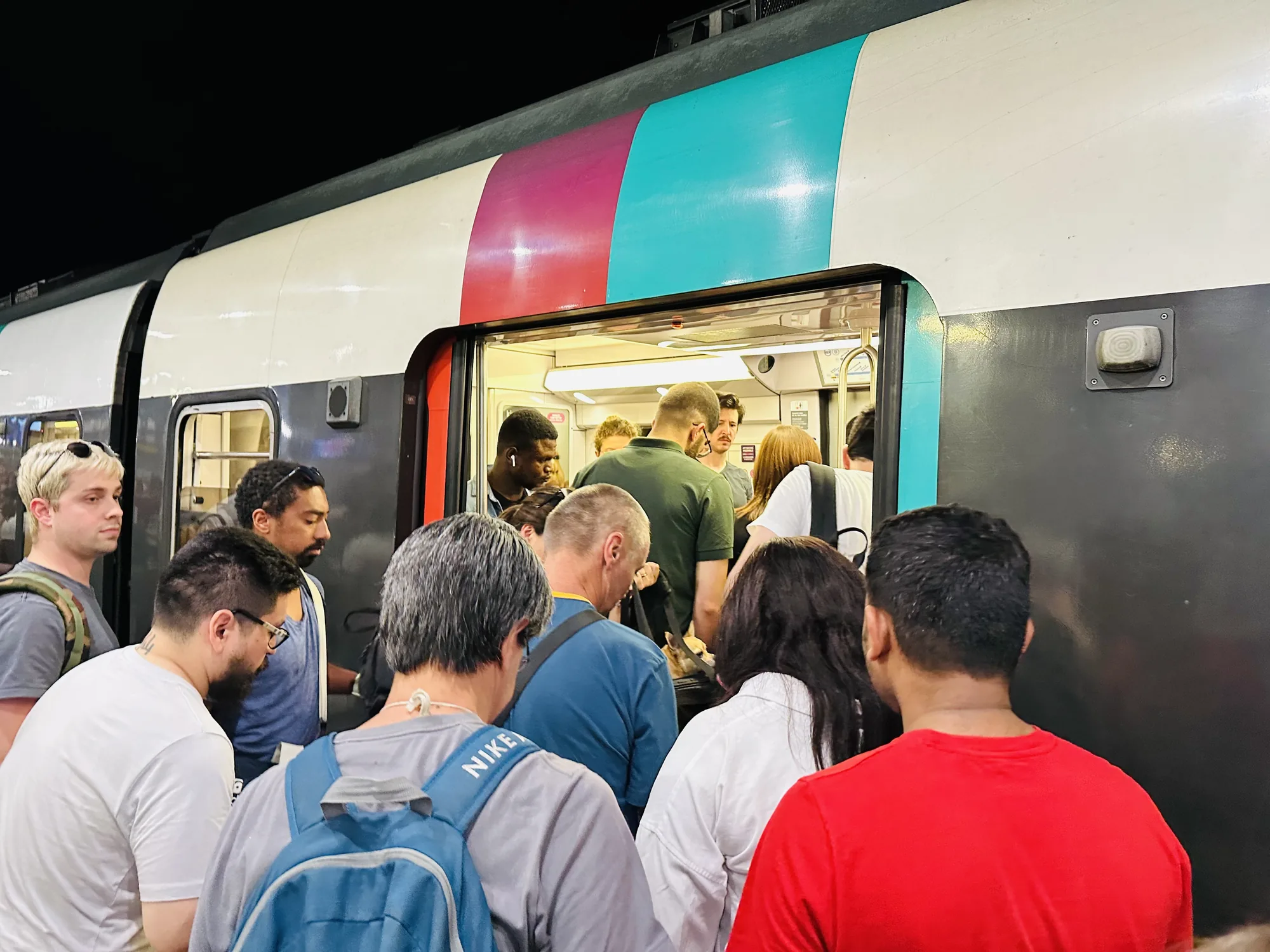 写真はフランス、パリで撮影されたRER（地域圏急行鉄道）の駅の様子を捉えています。夜間の駅のホームで、RERの車両に乗車しようとする多くの人々が写っています。人々は列車のドアから乗り込もうとしており、車内は混雑している様子が伺えます。様々な人種の人がいて、カジュアルな服装をしています。青色のNIKEのリュックを背負っている人もいます。車両の外観は白を基調とし、ピンクと水色のラインが入っています。