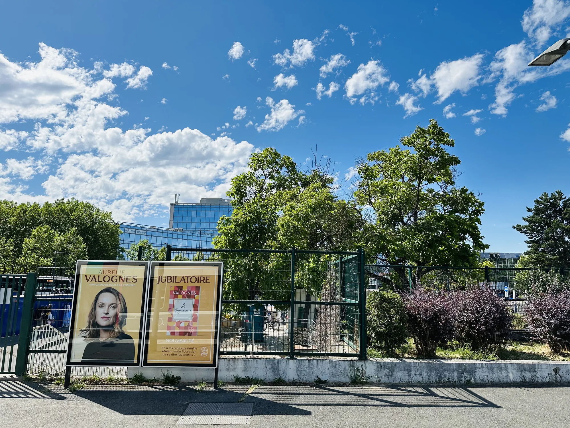 写真はフランスのパリで撮影されたもので、晴れた日の街並みが写っています。青空には白い雲が浮かび、明るい日差しが降り注いでいます。前景には、道路に面したフェンスがあり、その手前に2つの広告看板が設置されています。看板にはフランス語で書かれた映画か本の広告が貼られており、著名人の顔写真も見られます。フェンスの向こうには緑豊かな木々が生い茂り、背景にはガラス張りの近代的な建物が垣間見えます。全体的に明るく爽やかな雰囲気で、パリの日常風景を切り取ったような印象を受けます。