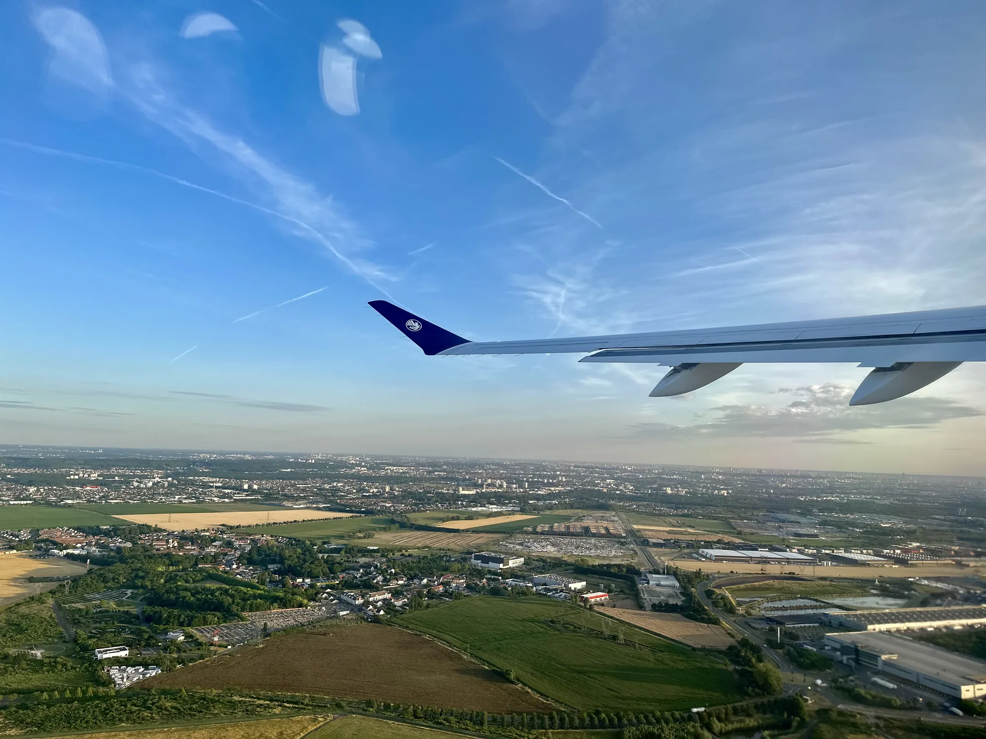 この写真は、フランスのパリ上空を飛行中の飛行機から撮影されたものです。機体の翼の一部が画面右上に写っており、翼の先端には航空会社のロゴが見えます。空は澄み渡っており、飛行機雲がいくつか見られます。地平線にはパリの街並みが広がっており、建物、緑地、道路などが確認できます。全体的に、美しい風景と飛行機の旅のワクワク感が伝わってくる写真です。