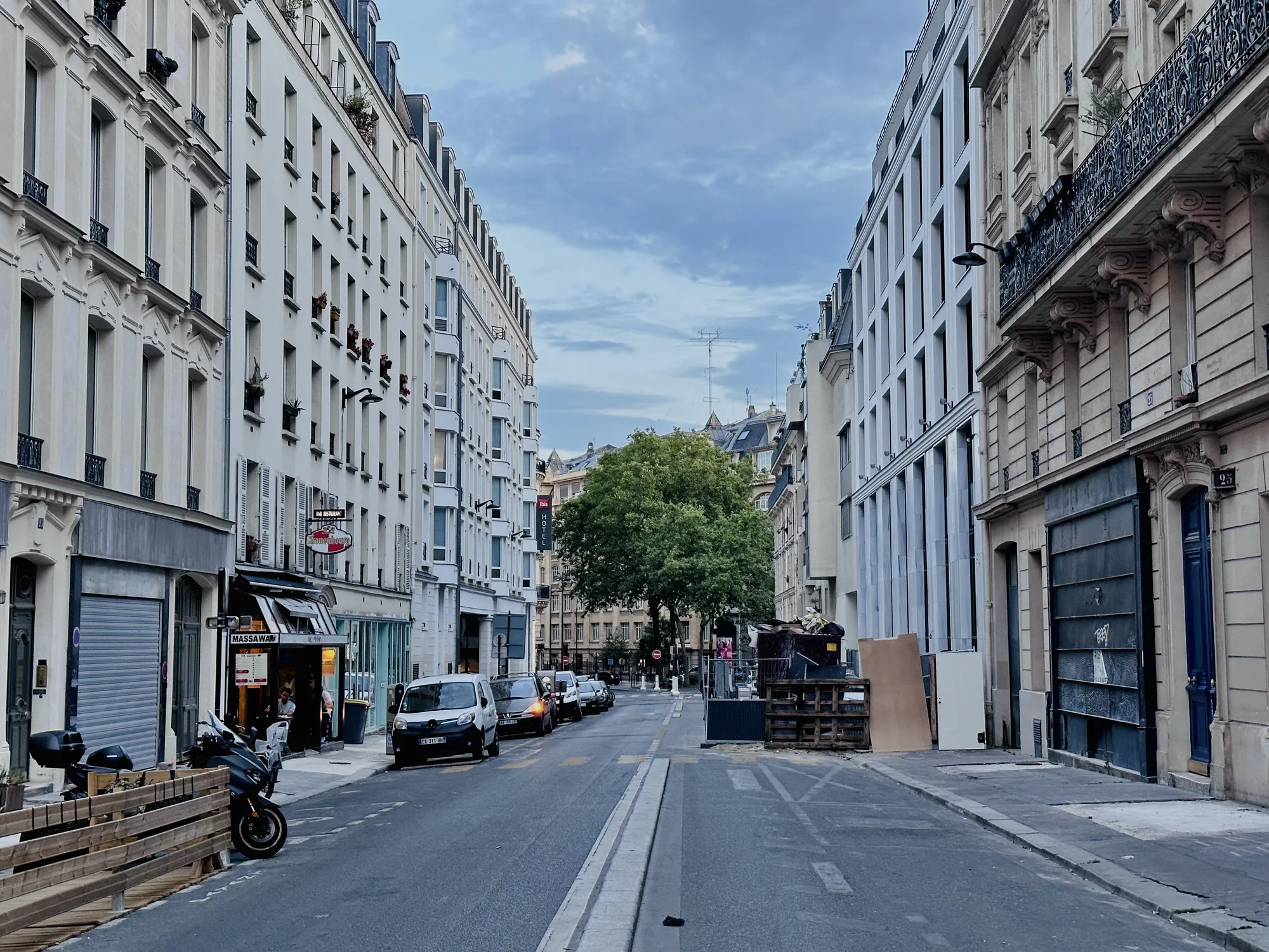フランスのパリで撮影された写真です。通りの奥には緑豊かな木々が見え、空は曇りがちです。通りの両側には、クラシックなパリの建築様式の建物が並んでいます。建物は白を基調としており、多くの窓やバルコニーが見られます。一部の建物には、窓辺に花が飾られています。通りの左側にはレストランやお店がいくつかあり、バイクが停まっています。通りには車が駐車しており、工事の資材のようなものが置かれています。全体的に、落ち着いた雰囲気で、パリの日常風景が捉えられています。