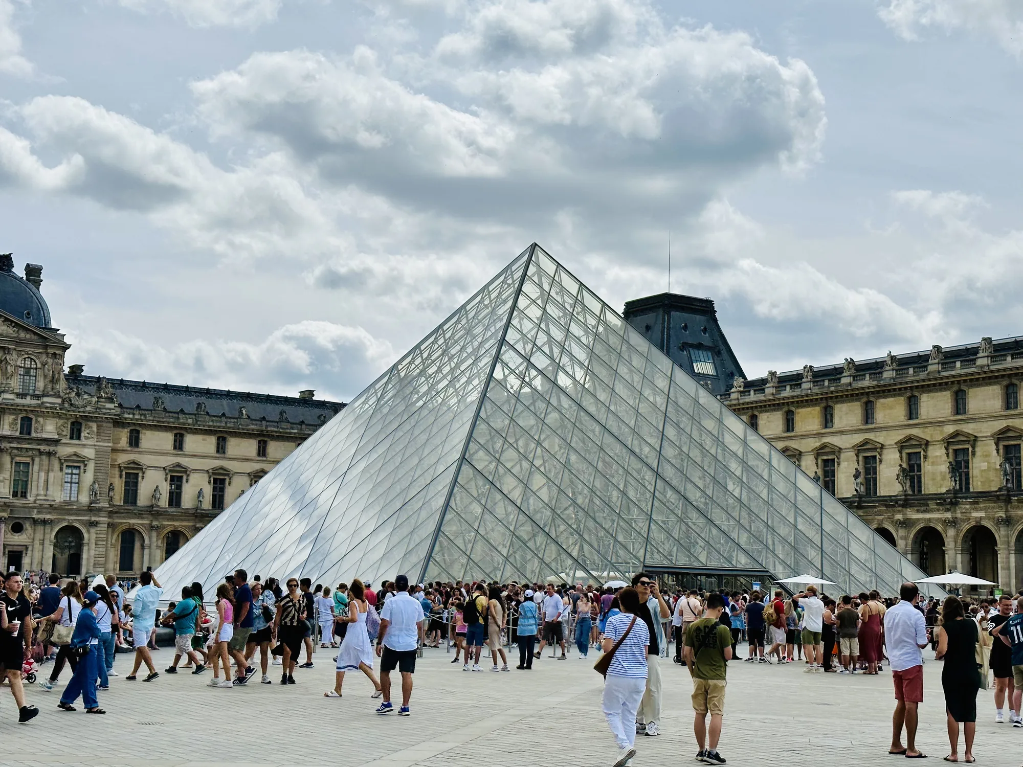 写真はフランスのパリにあるルーブル美術館で撮影されたものです。中心には、有名なガラスのピラミッドがそびえ立っています。ピラミッドの周囲には多くの観光客が集まっており、さまざまな服装の人々が行き交っています。ピラミッドの背景には、ルーブル美術館の古典的な建物の一部が見えます。空は少し曇っており、雲が広がっていますが、全体的に明るい光が感じられます。写真は、ルーブル美術館の活気ある雰囲気を捉えています。