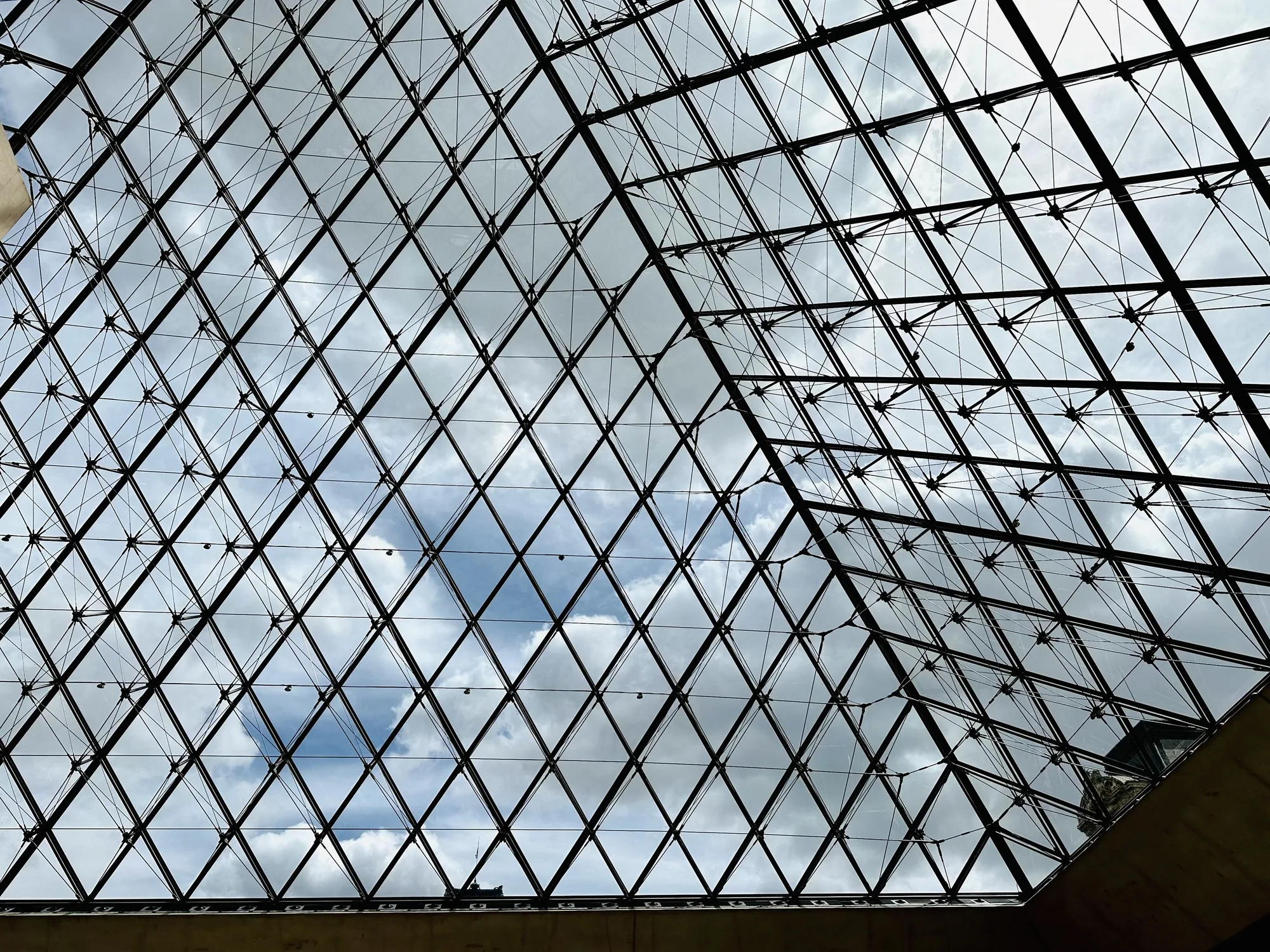 写真は、フランスのパリにあるルーブル美術館のピラミッドの内部から空を見上げた様子を捉えています。ピラミッドはガラスと金属製のフレームで構成されており、無数の菱形のガラス板が規則正しく配置されています。フレームは黒く、ガラスを通して見える空は、白っぽい雲が浮かぶ明るい空です。雲の隙間からは、青空がのぞいています。構造物の端の一部がわずかに写り込んでいます。全体的に、建築美と空の美しさが調和した、洗練された印象を与える写真です。