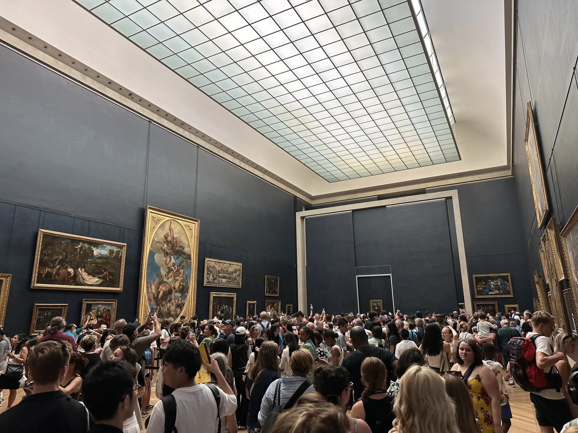この写真は、フランスのパリにあるルーブル美術館で撮影されたものです。巨大な展示室は、天井に格子状の明り取り窓があり、自然光が差し込んでいます。濃いグレーの壁面には、金色の額縁に入った絵画が多数展示されています。展示室は非常に混雑しており、多くの人々が絵画を鑑賞しようとしています。多くの人がスマートフォンを構えて写真を撮っており、その人気の高さが伺えます。絵画のジャンルは多岐にわたり、風景画や宗教画などが見られます。混雑ぶりから、特定の有名な作品の前に人が集まっている可能性があります。