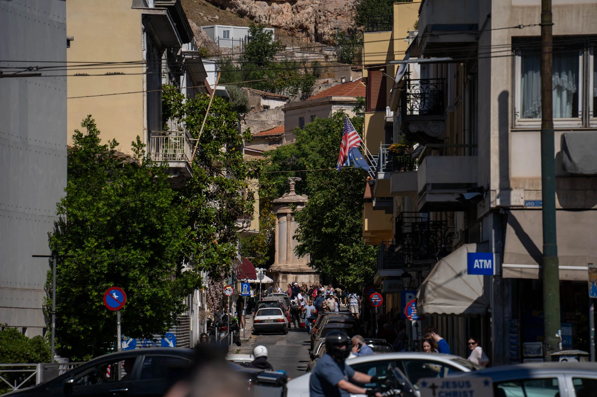 写真はギリシャのアテネで撮影されたもので、街並みを捉えています。通りの奥には古代の記念碑のような石造りの構造物があり、その周辺には多くの人々が集まっている様子が伺えます。通りの両側には建物が立ち並び、一部の建物にはバルコニーやアメリカ国旗とEUの旗が掲げられています。手前には車やバイクが通行しており、都市の喧騒が感じられます。全体的に日中の明るい光が差し込み、活気ある雰囲気です。ATMの看板や標識も見え、日常生活の一部が垣間見えます。