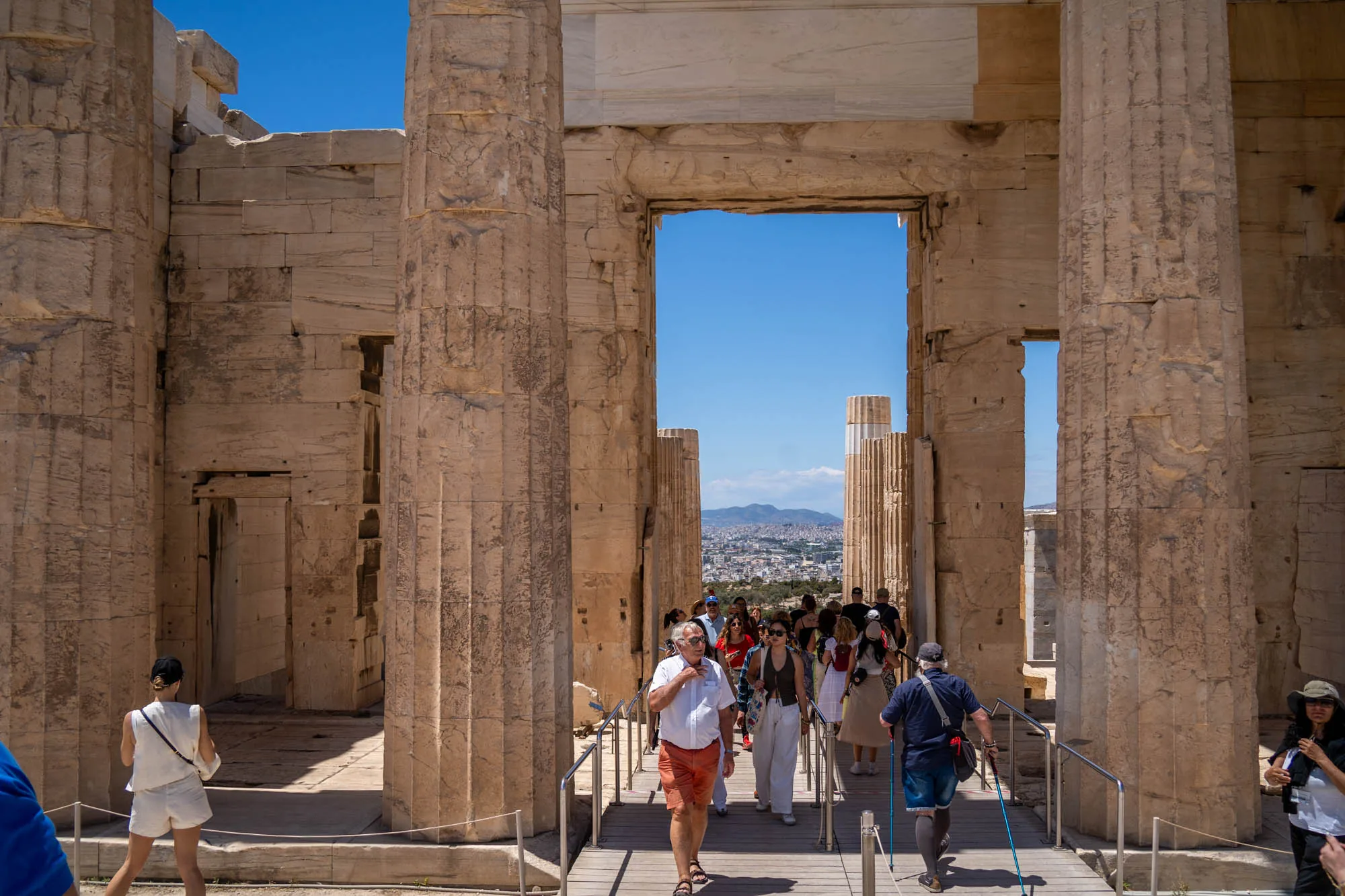 写真はギリシャのアテネで撮影されたものです。古代遺跡の一部が写っており、石造りの柱と壁で構成された構造物が印象的です。柱は風化しており、歴史を感じさせます。構造物の奥には、アテネの市街地が広がっており、遠くには山々も見えます。  遺跡内は観光客で賑わっており、様々な服装の人々が通路を歩いています。明るい日差しが降り注ぎ、開放的な雰囲気です。遺跡の壮大さと観光客の活気が組み合わさった、アテネの歴史と文化を感じさせる一枚です。