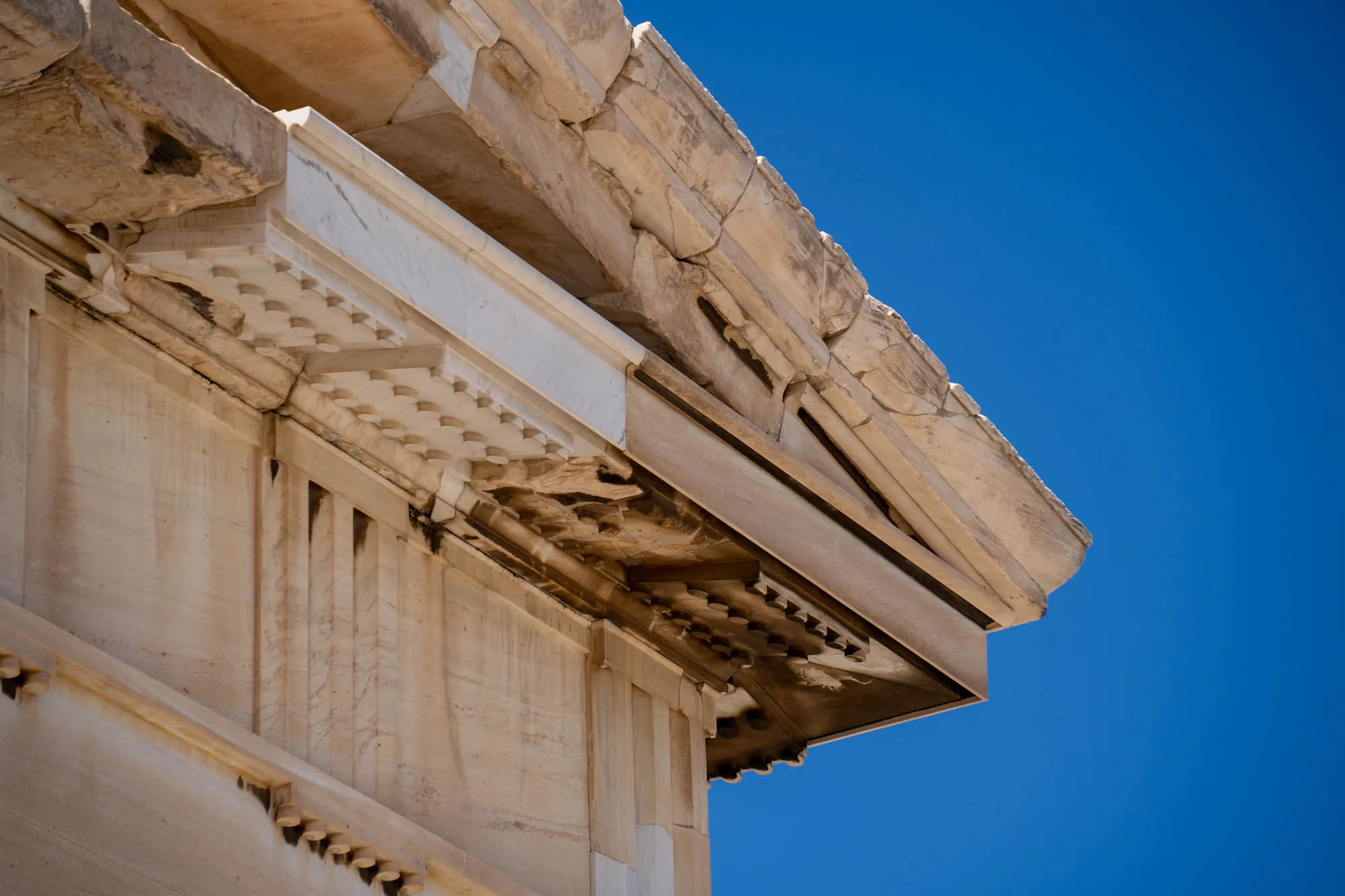 この写真はギリシャのアテネで撮影されたもので、古代建築の一部を捉えています。画面の大部分を占めるのは、精巧な装飾が施された石造りの構造物です。石材は長年の風化により、表面がざらつき、ひび割れも見られますが、その質感は歴史の重みを感じさせます。特に目を引くのは、規則的に配置された半円形の装飾や、垂直方向に伸びる溝状のデザインです。これらの装飾は、光と影のコントラストを生み出し、建築物の美しさを際立たせています。背景には、鮮やかな青空が広がっており、石造りの構造物とのコントラストが非常に印象的です。全体的に、写真からは古代ギリシャの建築技術の高さと、悠久の時を経た建造物の荘厳さが伝わってきます。