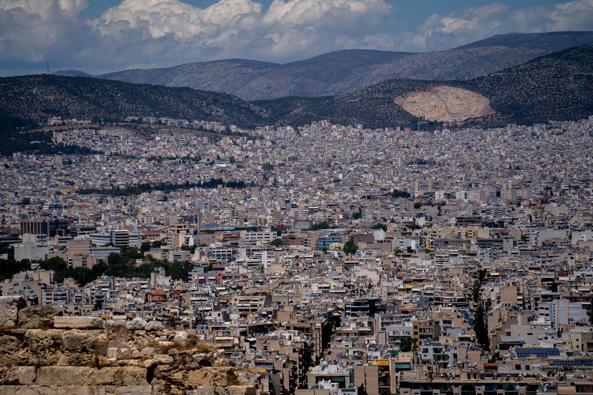 写真はギリシャのアテネで撮影された都市景観です。前景には古代の石壁の一部が写っており、そこから視線は広大な市街地へと広がります。建物が密集しており、そのほとんどが白やベージュの色調で統一されています。建物の高さは様々ですが、全体として均質な印象を与えます。市街地の背景には山々が連なり、山頂付近には雲が浮かんでいます。山の斜面には木々が点在していますが、一部には採石場のような露出した岩肌も見られます。空は晴れており、明るい日光が都市全体を照らしています。全体的に、歴史的な景観と現代の都市生活が融合した印象を受ける写真です。