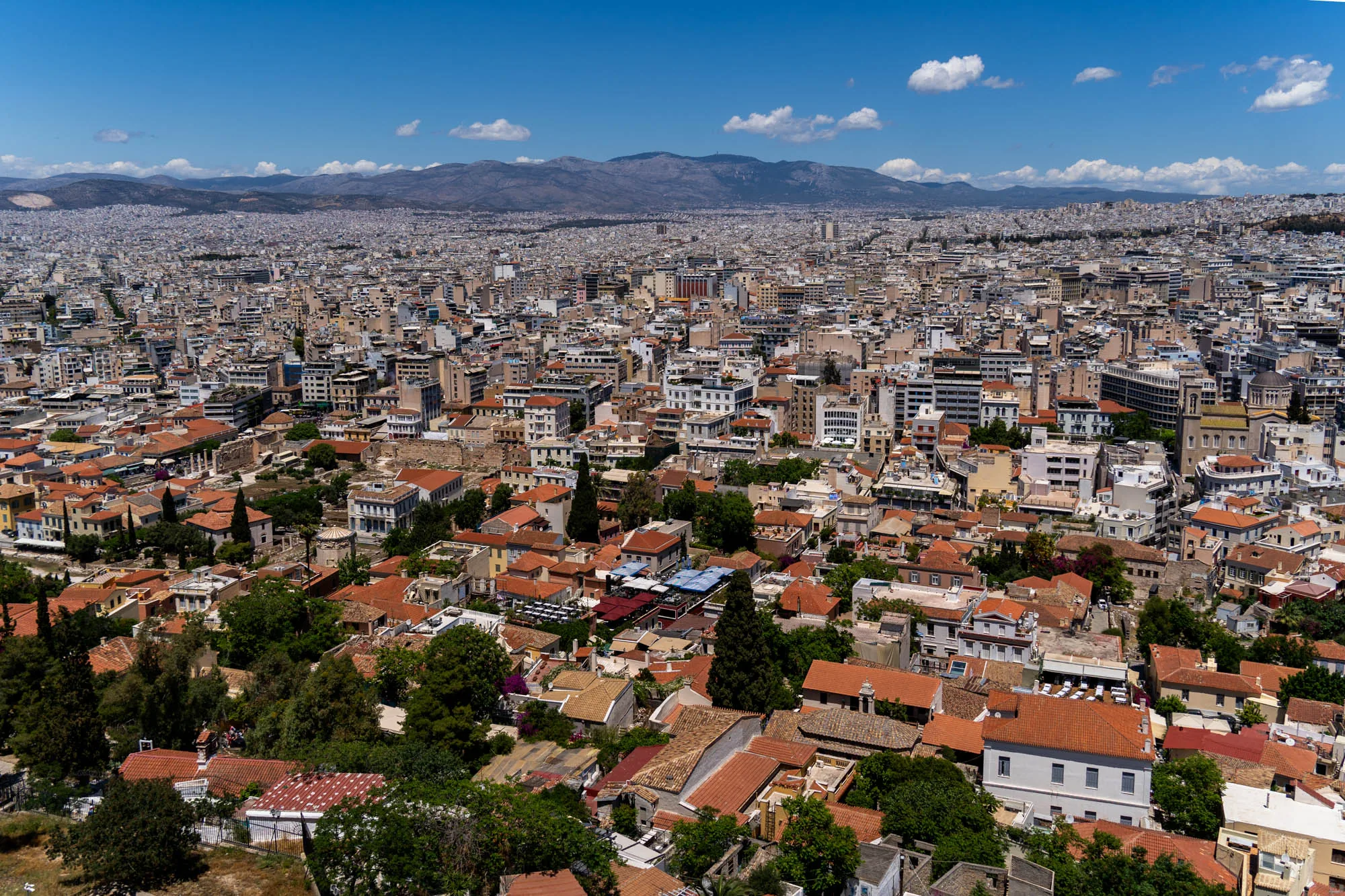 写真はギリシャのアテネで撮影された、高所からの都市景観です。手前には赤茶色の屋根が連なる住宅街が広がり、緑豊かな木々が点在しています。遠景には、建物が密集した市街地が地平線まで続いており、その奥には山々がそびえ立っています。空は青く、白い雲が浮かんでおり、全体的に明るく開放的な雰囲気です。建物の種類は様々で、新旧の建物が混在している様子が伺えます。太陽の光が建物の壁面に当たり、陰影を作っているため、奥行きが感じられます。遠くの山々にかかる霞が、風景全体に奥行きと広がりを与えています。