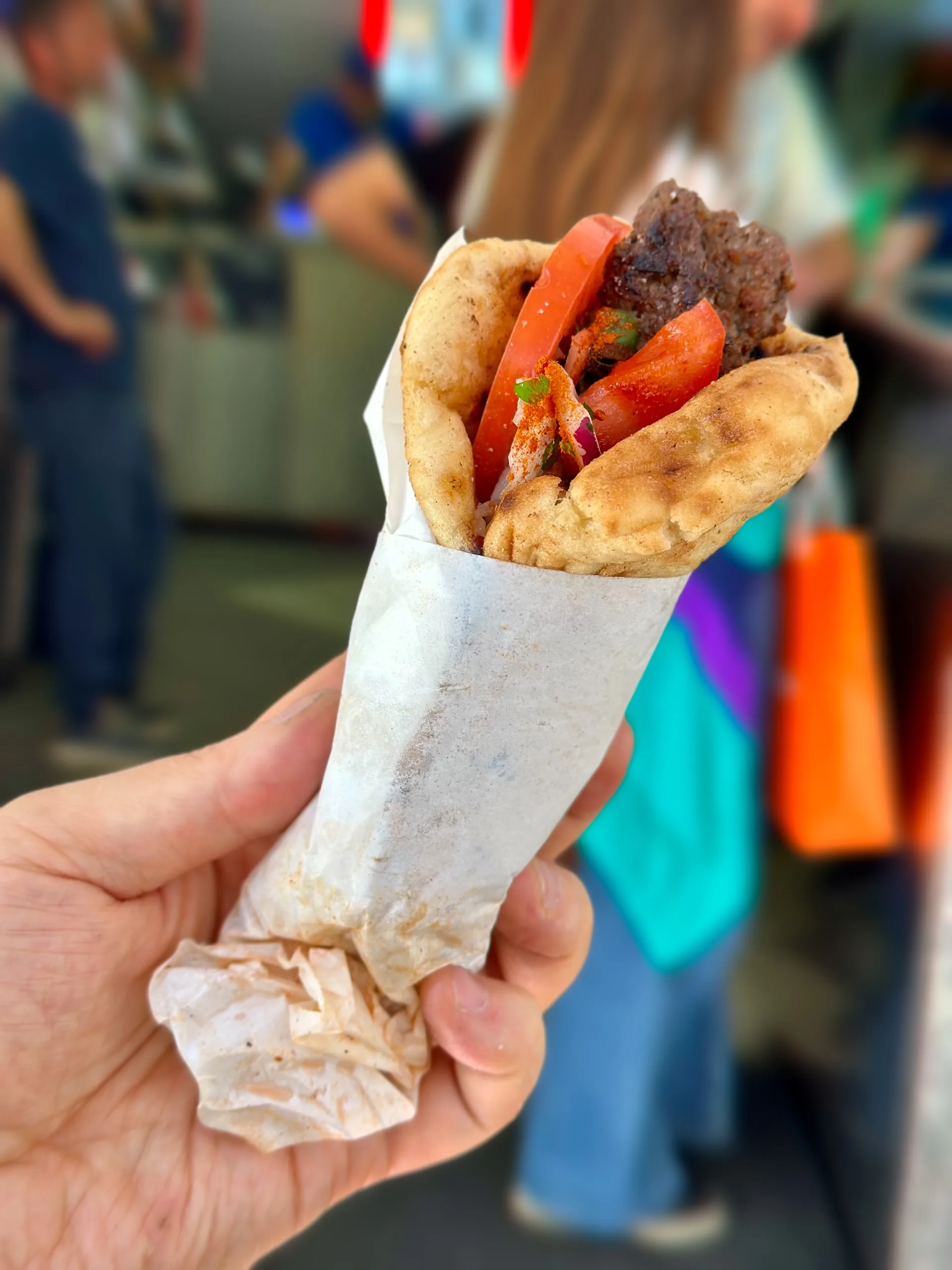 写真には、ギリシャのアテネで撮影されたピタサンドが写っています。ピタは白い紙に包まれており、人の手に持たれています。ピタの中には、焼かれた肉、トマト、タマネギなどの具材が入っています。背景には、ぼやけた人々が写っており、屋台のような場所であることがわかります。全体的に、ローカルな雰囲気の漂う、美味しそうなストリートフードの写真です。