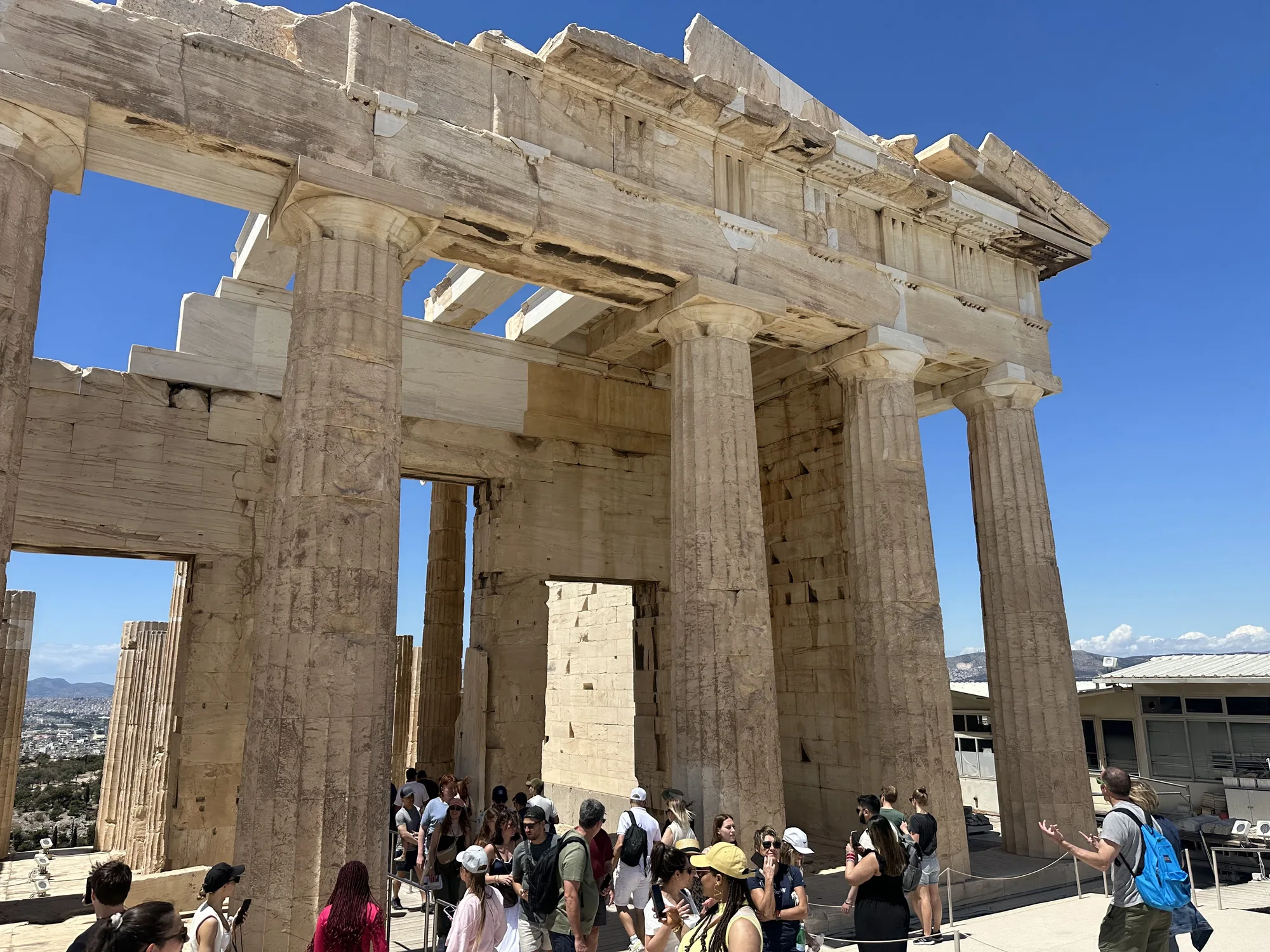 写真はギリシャのアテネで撮影されたもので、パルテノン神殿の一部を捉えています。青い空を背景に、巨大な石柱と構造物が堂々とそびえ立っています。石材はベージュ色で、長い年月を経て風化した様子が見て取れます。柱の彫刻や装飾は細部まで丁寧に作り込まれており、古代ギリシャの高度な建築技術を物語っています。  写真の下部には多くの観光客が写っており、その規模の大きさを際立たせています。人々はガイドの説明を聞いたり、写真を撮ったりしながら、この歴史的な建造物を楽しんでいるようです。建物の隙間からは、アテネの街並みが遠くに見え、壮大な景観が広がっています。全体的に、歴史の重みと観光地の賑わいが感じられる一枚です。