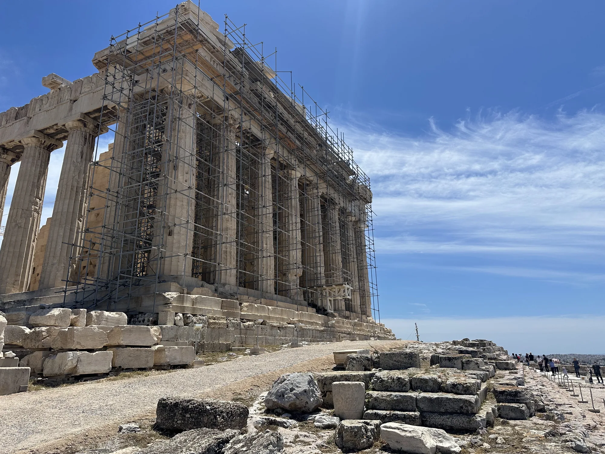 写真はギリシャのアテネにあるパルテノン神殿を捉えたものです。青い空を背景に、荘厳な神殿の一部が写っています。神殿は修復作業中のようで、一部が足場で覆われています。巨大な石柱が整然と並び、古代の建築技術の高さがうかがえます。神殿の基部には、崩れた石材が積み上げられており、長い年月を経た遺跡の風化を感じさせます。手前には、神殿へと続く砂利道と、石畳のような場所があり、観光客が訪れている様子が伺えます。空は晴れ渡り、薄い雲が流れています。全体的に、歴史的な建造物の威厳と、修復作業の様子が伝わる写真です。