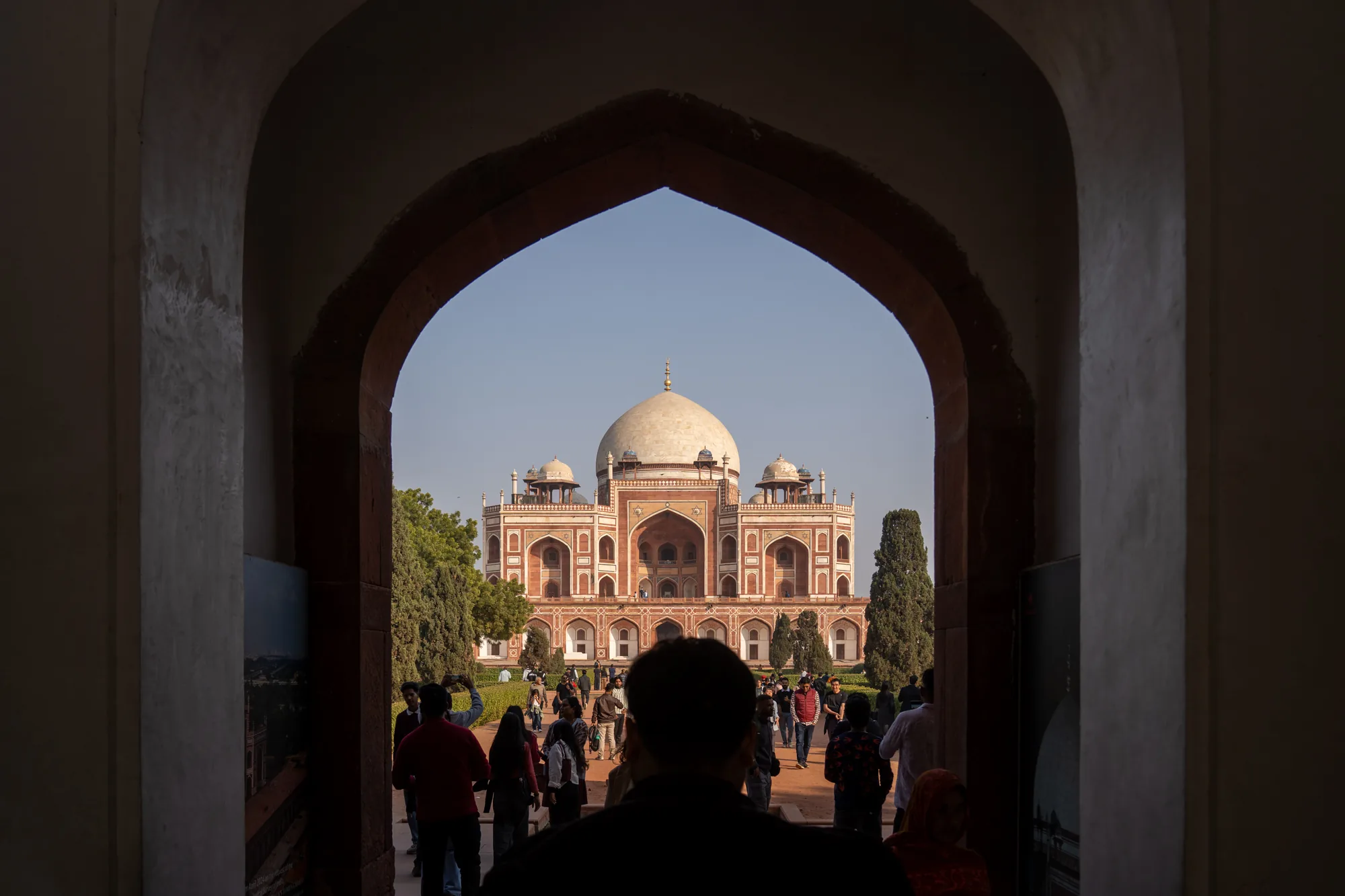 インドのニューデリーで撮影されたフマユーン廟の写真です。写真はアーチ型の通路から撮影されており、通路の縁は赤茶色で、背景の青空によく映えています。通路の前には多くの人が歩いており、観光客と思われます。フマユーン廟は、赤砂岩と白い大理石で建てられた壮麗な建物で、大きなドームが特徴です。周囲には緑豊かな庭園が広がっており、景観を美しく彩っています。写真全体は、歴史的な建造物の荘厳さと、それを訪れる人々の活気が組み合わさった印象を与えます。