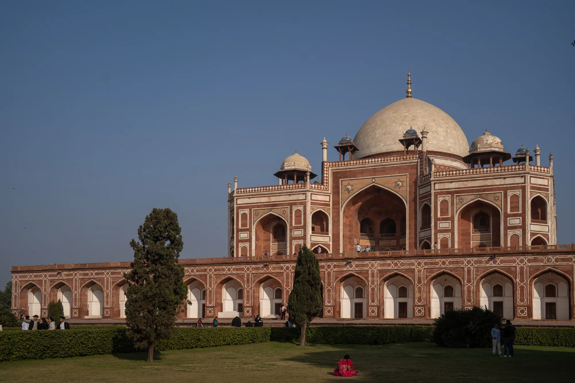 写真は、インドのニューデリーにあるフマユーン廟を捉えています。青空を背景に、赤茶色と白を基調とした壮麗な建物が堂々とそびえ立っています。建物の特徴的な大きなドーム型の屋根は、薄いクリーム色をしており、頂上には金色の装飾が施されています。建物全体は、アーチ型の窓や幾何学的な模様が施された壁面で飾られており、精緻な装飾が施されています。  建物の手前には、緑豊かな芝生の庭が広がっており、数本の木々が植えられています。芝生の上や建物の周りには、観光客と思われる人々が散見され、ゆったりとした雰囲気が漂っています。鮮やかな赤いサリーをまとった女性が芝生に座っており、印象的なアクセントになっています。全体的に、歴史的な建造物と自然が調和した、美しい光景です。