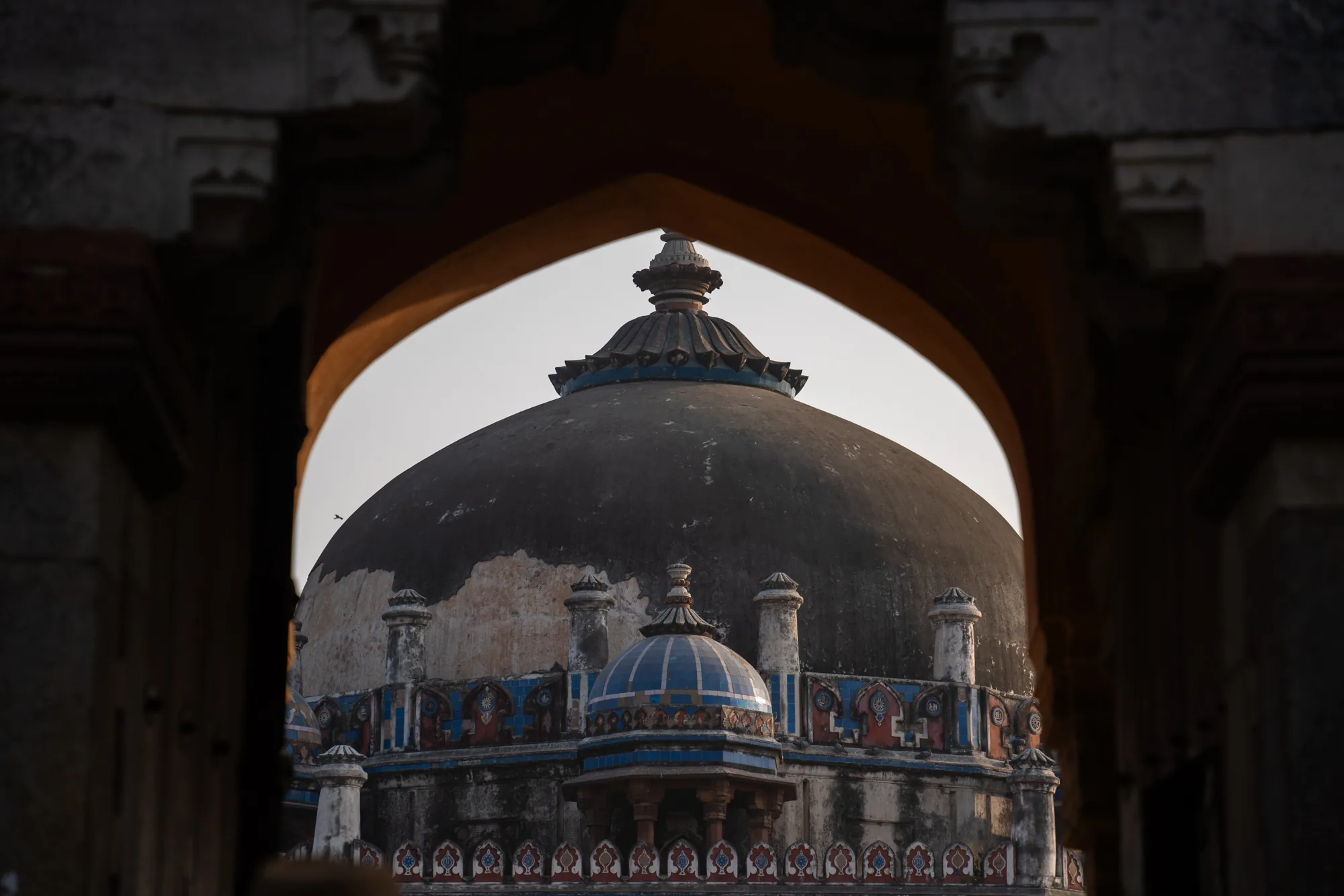 この写真はインドのニューデリーで撮影されたもので、石造りのアーチ越しに見えるドーム建築が印象的です。アーチは背景を額縁のように切り取り、ドームの存在感を際立たせています。ドームは大きく、灰色がかった色合いで、表面には経年によるシミや剥がれが見られます。その下には、青色を基調とした装飾が施された小さなドームが配置されており、全体として複雑で美しいデザインとなっています。さらに下には、花柄のような模様が繰り返し描かれた装飾があり、細部にまでこだわりが感じられます。写真は、歴史的な建造物の静寂と威厳を伝えており、インドの文化的な豊かさを感じさせる一枚です。