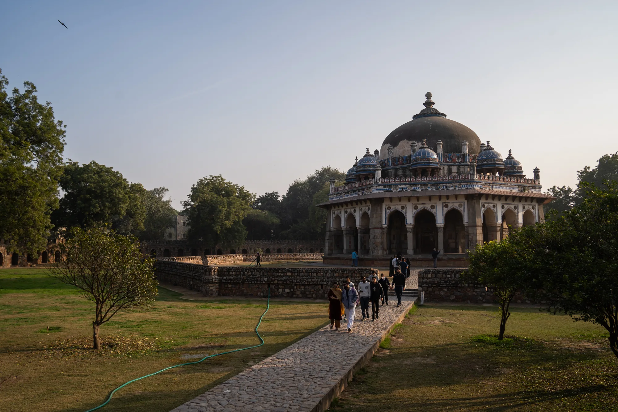 写真はインドのニューデリーで撮影されたもので、歴史的な庭園と廟が写っています。画面中央には、ドーム型の屋根を持つ美しい廟がそびえ立っています。廟の周囲にはアーチ状の構造が並び、青や白の装飾が施されています。石畳の道が廟へと続いており、数人の人々がそこを歩いています。庭園は広々としていて、緑豊かな芝生や木々が生い茂っています。画面左奥には石造りの壁が見え、さらに奥には別の建物の一部が確認できます。空は晴れており、鳥が1羽飛んでいます。全体的に、歴史的な建造物と自然が調和した、静かで美しい風景です。観光客と思しき人々が、この歴史的な場所を訪れている様子が伺えます。