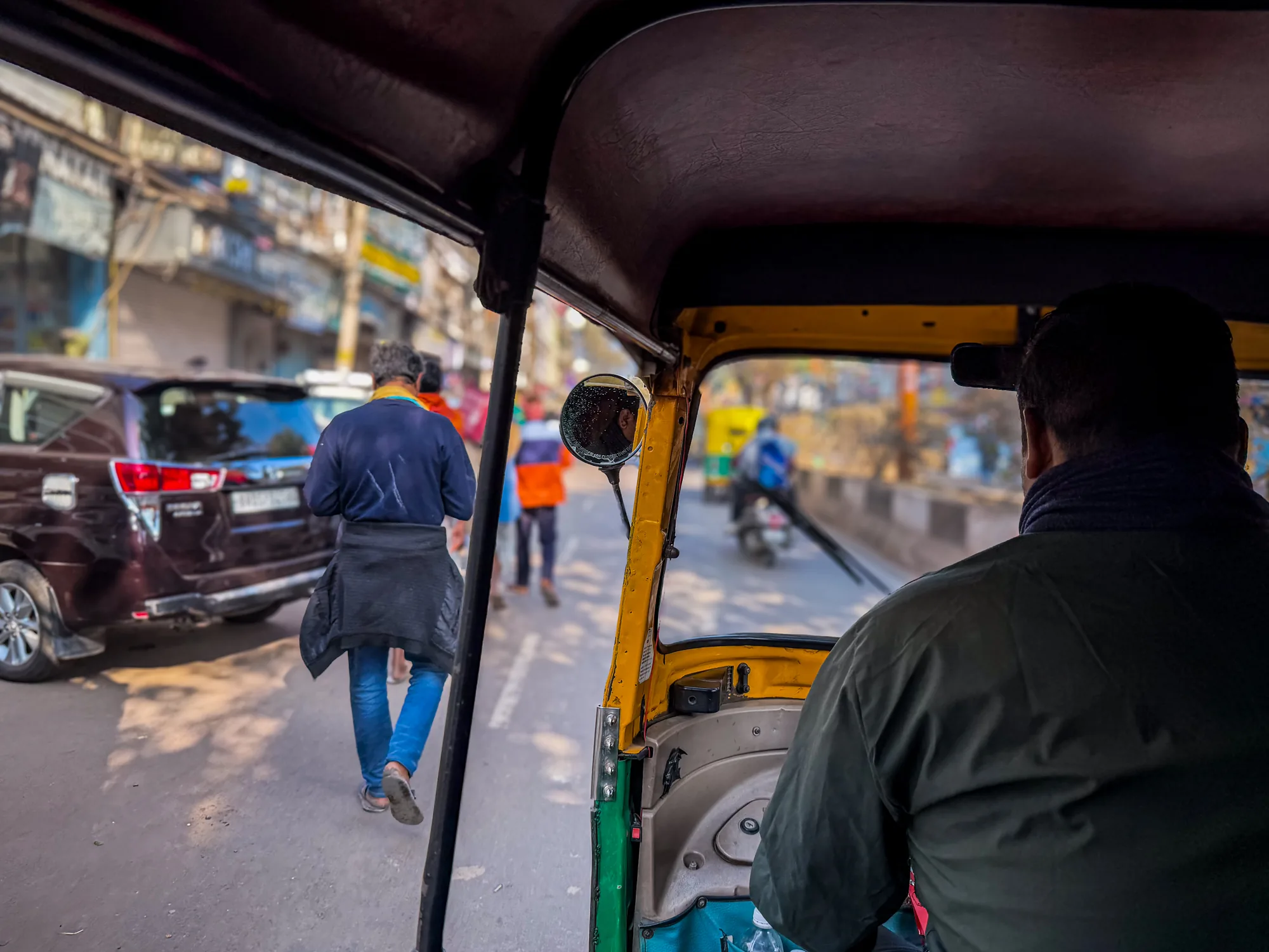 写真は、インドのニューデリーで撮影されたオートリクシャーの車内からの視点です。オートリクシャーの運転手の後ろ姿が写っており、彼はダークグリーンのシャツを着ています。運転手の肩越しには、フロントガラス越しに見えるデリーの街並みが広がっています。路上には歩行者や他の車両が見え、活気ある雰囲気が伝わってきます。画面左側には、男性が紺色のシャツを着ており、上着を腰に巻いて歩いています。その隣には自動車が止まっており、背景には建物が並んでいます。オートリクシャーの内部は黄色と緑色で塗られており、旅行の雰囲気とインドの独特な風景を感じさせる一枚です。