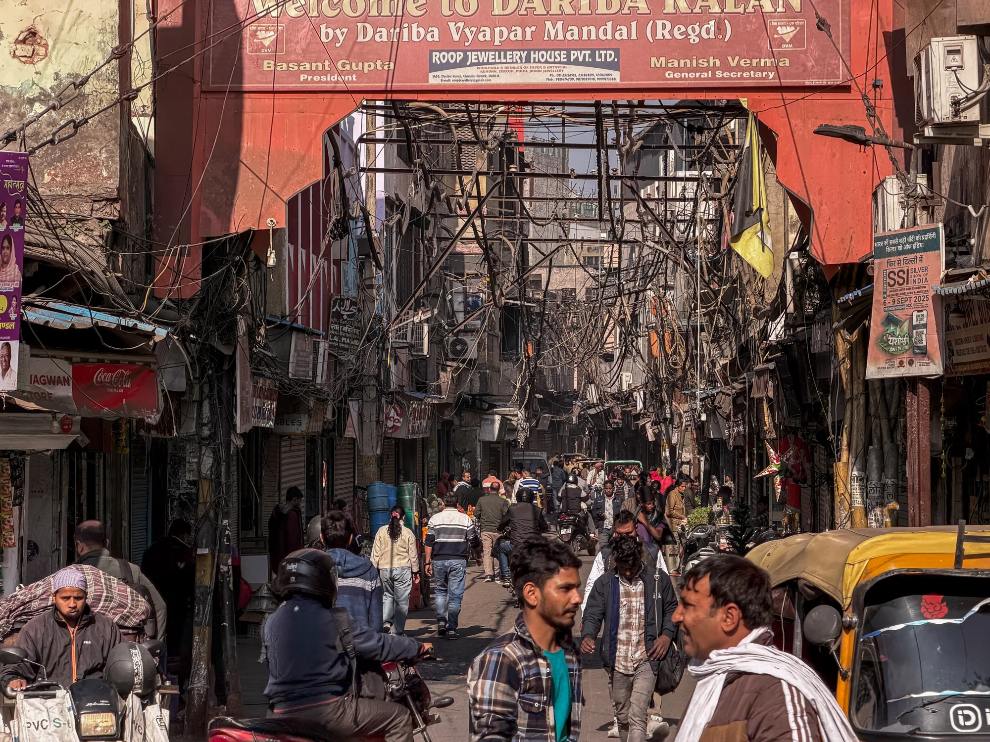 インドのニューデリーで撮影された写真です。写真には、活気に満ちた通りと、その通りを行き交う人々が写っています。通りは「Welcome to DARIDA KALAN by Dariba Vyapar Mandal (Regd.)」と書かれた赤いアーチで飾られています。建物の間には無数の電線が張り巡らされ、複雑な模様を描いています。通り沿いには様々なお店が並び、コカコーラの看板や広告、そしてヒンディー語の文字が見られます。人々は徒歩やバイク、オートリクシャーで移動しており、街の活気を伝えています。特にオートリクシャーは鮮やかな黄色で、画面の右側に大きく配置されています。全体的に、この写真は混沌とエネルギーに満ちたインドの都市生活の一面を捉えています。