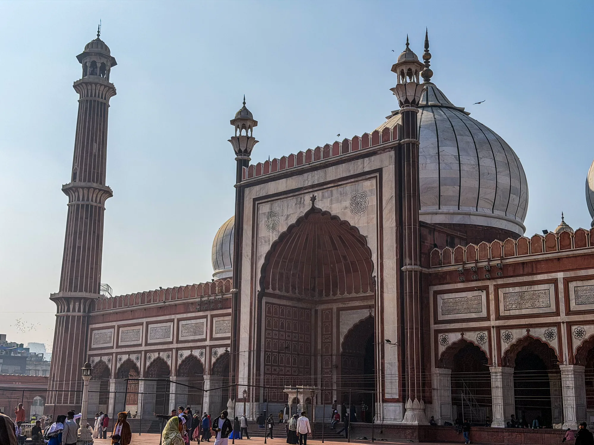 写真はインドのニューデリーにあるモスクを捉えたものです。空は晴れ渡り、建物全体を明るく照らしています。画面左側には高くそびえ立つミナレット（尖塔）があり、赤茶色の石造りで垂直方向に模様が刻まれています。ミナレットの頂上には小さなドーム状の構造物が見えます。モスクの本体は赤茶色と白の大理石でできており、巨大なドームが特徴的です。ドームは白く、縦方向にラインが入っています。建物にはアーチ型の入り口や装飾が施されており、壁面には文字や模様が刻まれています。建物の前には人々が歩いており、モスクの大きさを際立たせています。全体的に、荘厳で美しい雰囲気の写真です。