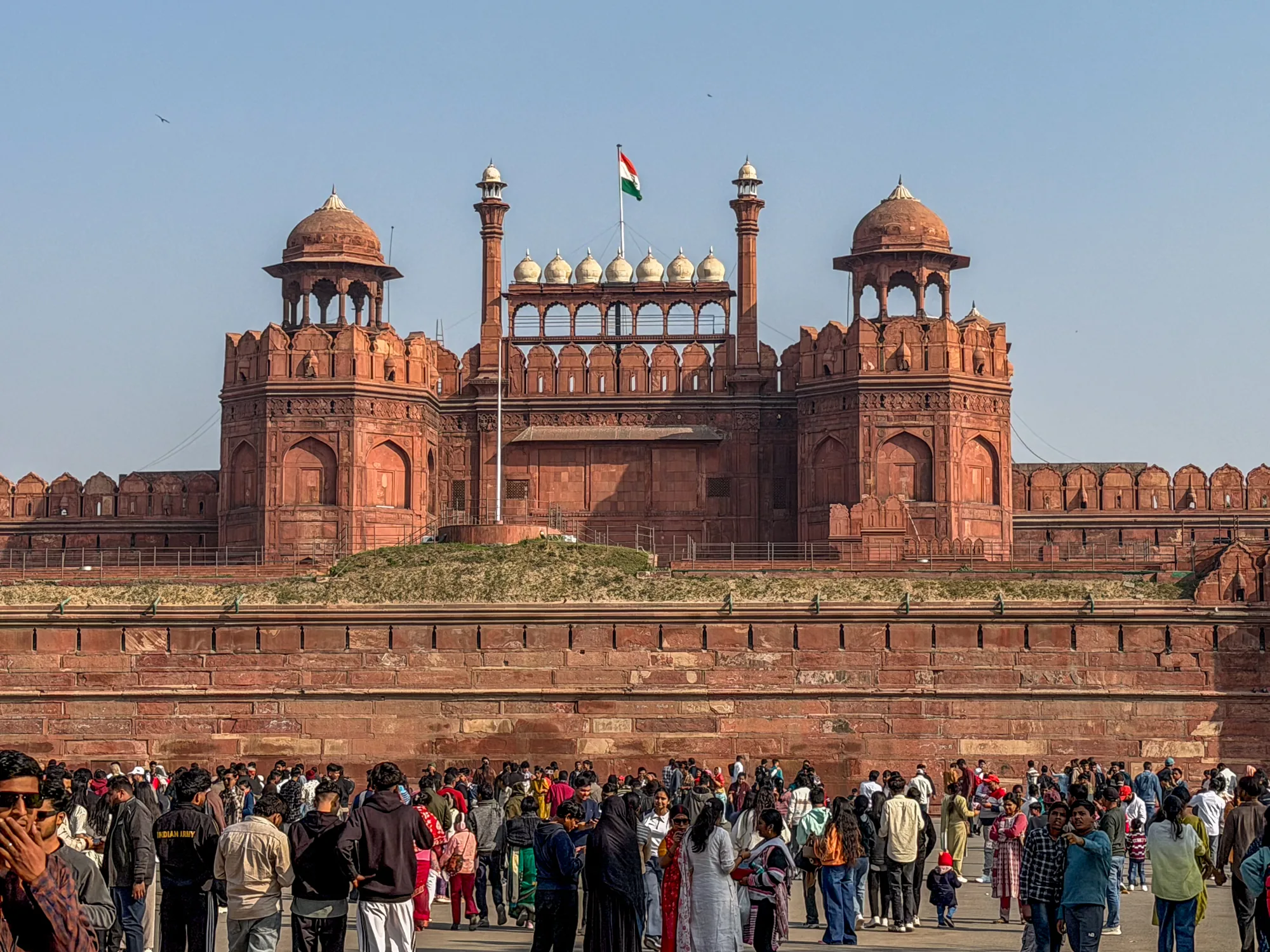 インドのニューデリーにあるラール・キラー（赤い砦）を撮影した写真です。鮮やかな赤い砂岩でできた壮大な砦が、写真全体を占めています。砦の上にはインドの国旗が掲げられています。砦の正面には、多くの人々がおり、観光客や地元の人々が混ざり合っているようです。様々な服装の人々がいて、活気ある雰囲気を感じさせます。空は晴れており、日中の撮影であることがわかります。全体的に、インドの歴史的建造物と人々の生活が組み合わさった、活気のある一枚です。