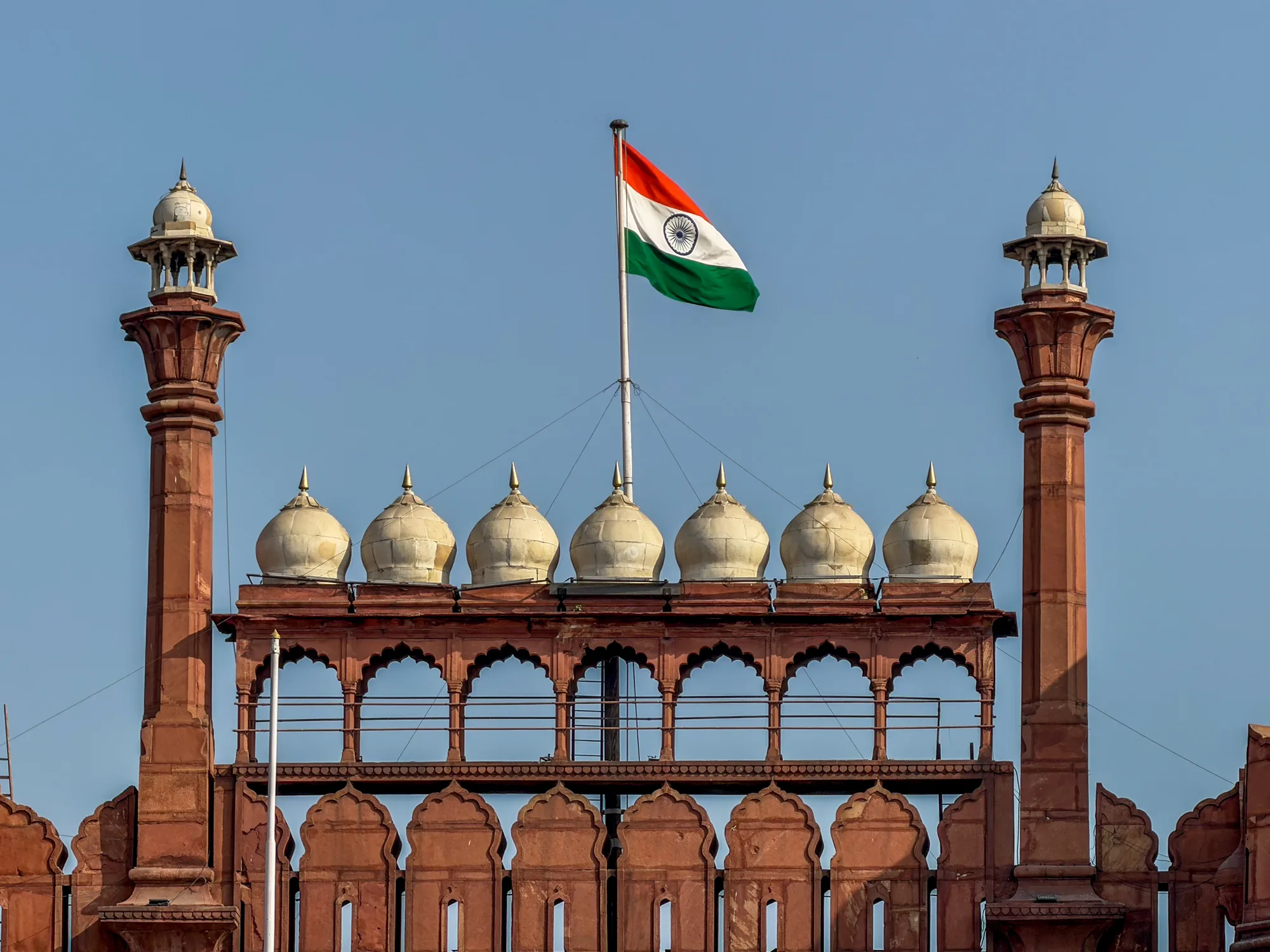 インドのニューデリーで撮影された写真です。画像には、赤い砂岩でできた印象的な建造物の一部が写っています。これは、おそらくデリーの赤い城（ラール・キラー）の一部でしょう。建物の上部には、丸いドームが連なっており、その上にインドの国旗が掲げられています。空は晴れ渡っており、建物の赤色と空の青色のコントラストが鮮やかです。建物の細部に至るまで丁寧に描写されており、歴史的建造物の重厚感と、国旗が象徴するインドの誇りが感じられます。建造物のアーチ型のデザインや、装飾的な要素が、さらに写真に深みを与えています。