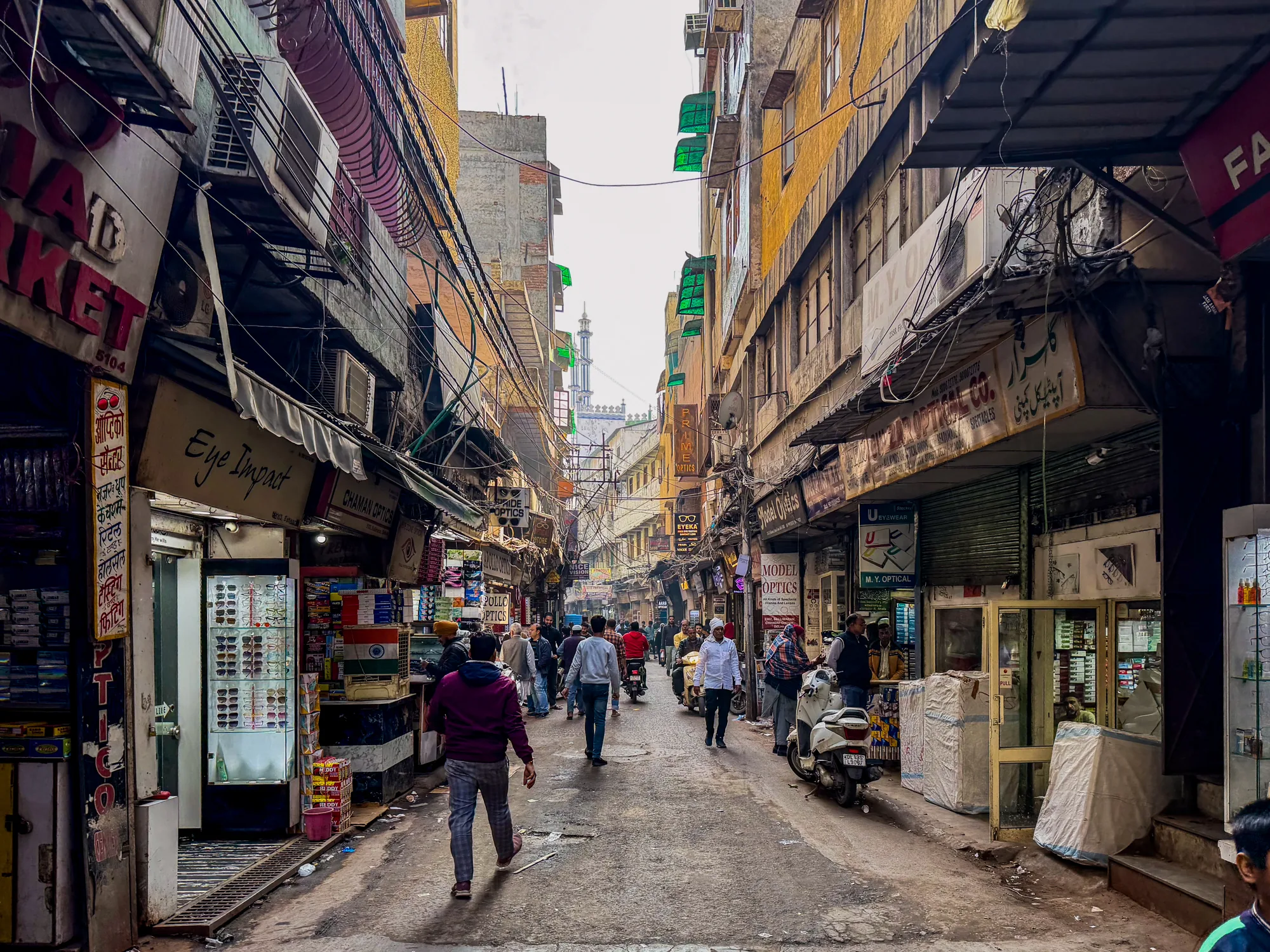 写真は、インドのニューデリーで撮影されたと思われる、活気のある商店街の様子を捉えています。狭い路地の両側には、様々な商品が並んだ商店が軒を連ね、多くの人々が行き交っています。店舗の看板には、英語とヒンディー語と思われる文字が混在しており、ローカルな雰囲気を醸し出しています。電線が複雑に張り巡らされ、建物に設置されたエアコンの室外機などが生活感を感じさせます。路地の奥にはモスクのミナレットの一部が見え、文化的背景を示唆しています。全体的に活気があり、雑多なエネルギーに満ちた、インドの都市の一面を切り取った写真です。多くの店がメガネ店であることが特徴的です。