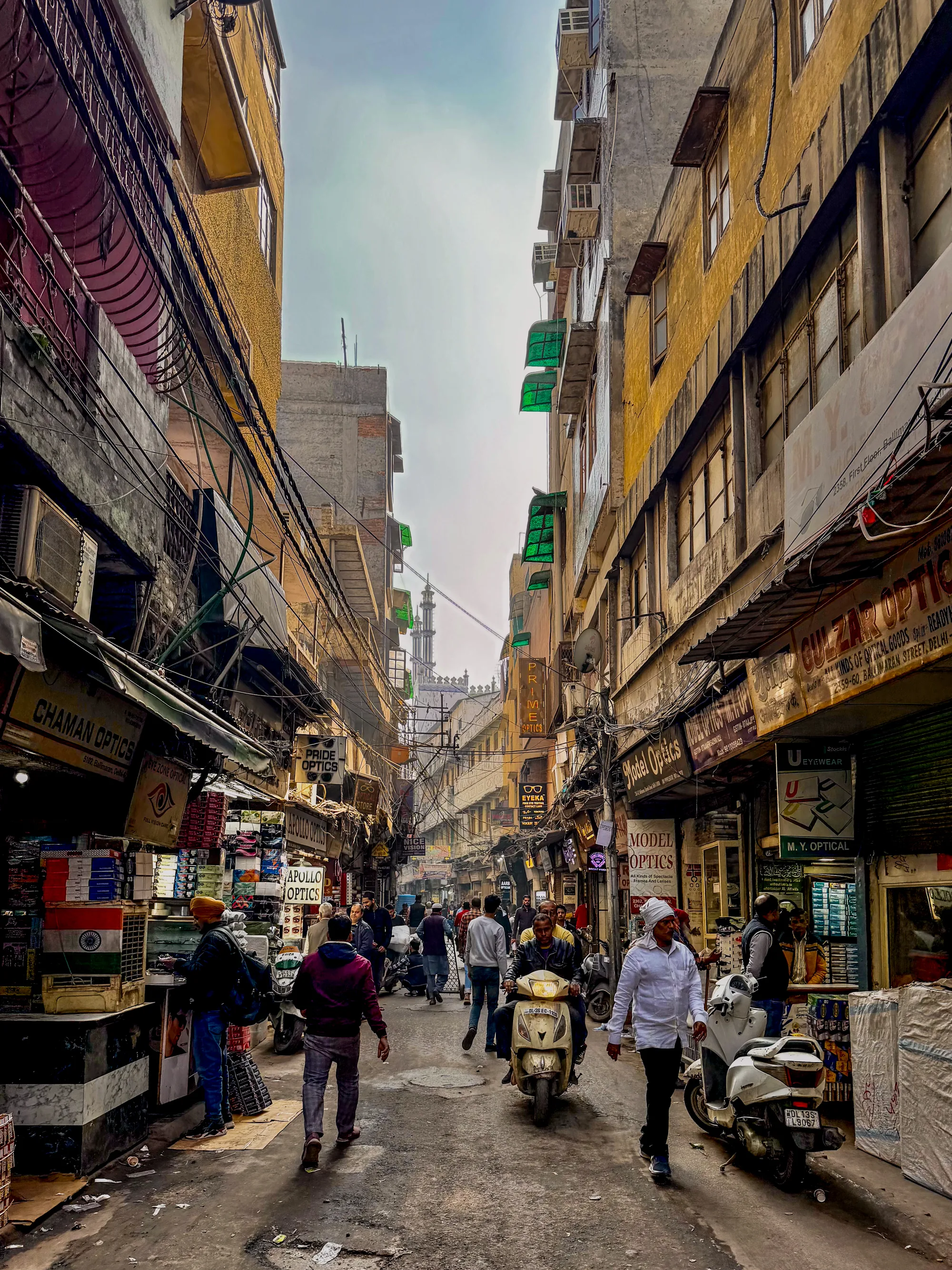 この写真はインド、ニューデリーの賑やかな通りを捉えています。狭い路地には多くの人々が行き交い、スクーターが走り抜けています。通りの両側には、多くの店が軒を連ねており、看板には「OPTICS」という文字が頻繁に見られます。これらの店は眼鏡店であることがわかります。店の外には商品が並べられ、活気に満ちた市場の雰囲気を醸し出しています。建物は古いレンガ造りで、エアコンの室外機や張り巡らされた電線が生活感を感じさせます。遠くにはモスクのミナレットが見え、街の風景に宗教的な要素を加えています。全体的に、混沌としていながらも活気に満ちた、典型的なインドの都市の風景が描写されています。