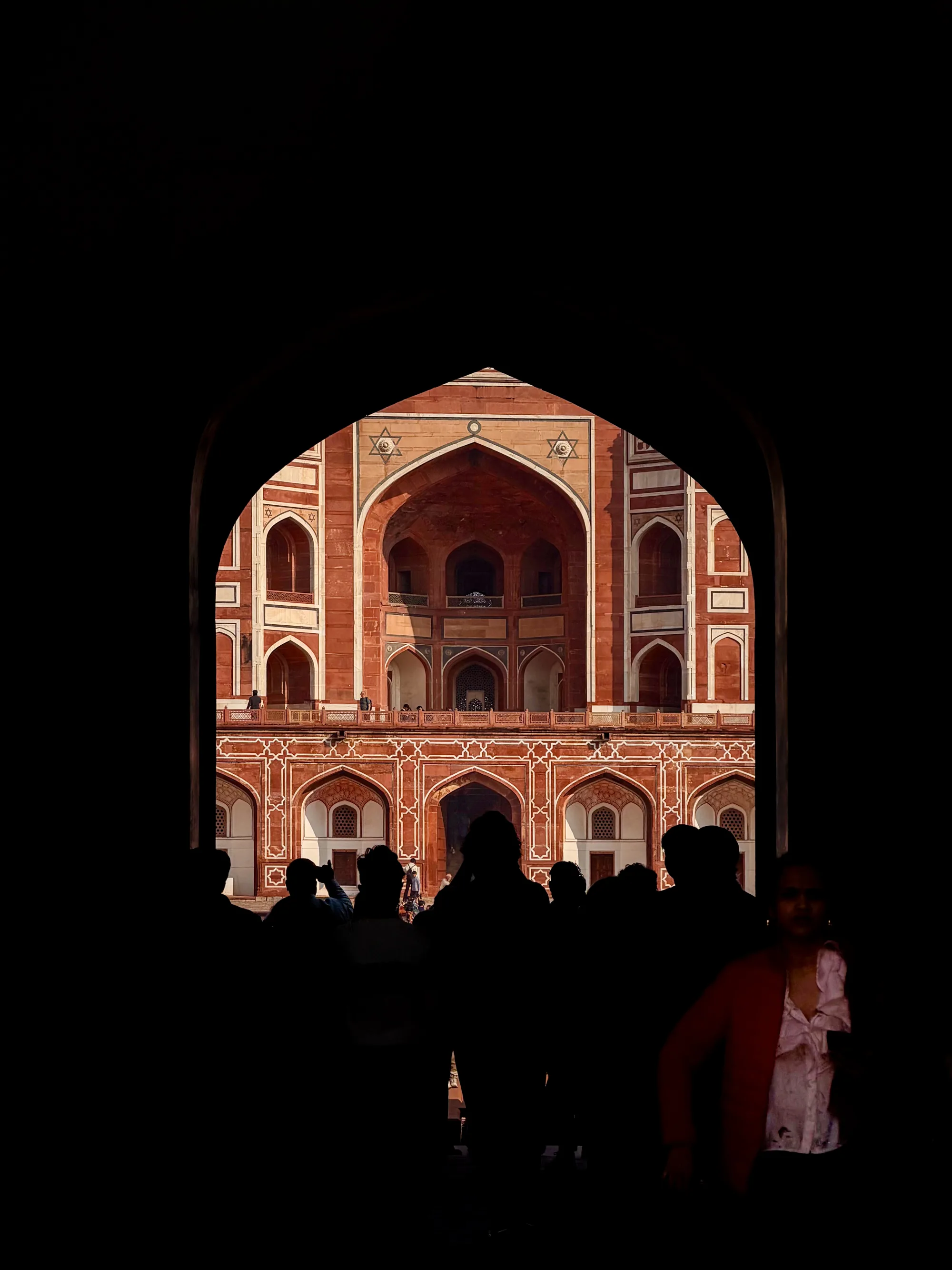 写真は、インドのニューデリーで撮影されたものです。手前の暗いアーチ状の開口部から、壮麗な建築物がフレームに収められています。建物の外壁は赤茶色を基調とし、白い装飾模様が施されています。アーチ型の窓やバルコニーが幾重にも重なり、その精緻なデザインが目を引きます。建物の上部には星形の装飾が施されています。  アーチの入り口には、多くの人々のシルエットが写っており、彼らは建築物を鑑賞しているようです。右側には、赤色のジャケットを着た女性がおり、彼女の顔がわずかに確認できます。写真全体はコントラストが強く、暗闇と光の対比が印象的です。建物の壮大さと、そこに集まる人々の様子が伝わってくる一枚です。