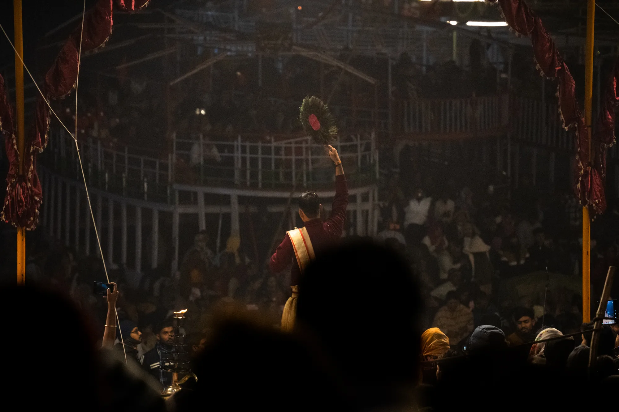 写真はインドのバラナシで撮影されたものと思われます。夜の儀式の様子が写されており、暗い中にも厳かな雰囲気が漂っています。正面にいる人物は、赤い衣装を身につけ、扇のようなものを持って腕を上げています。おそらく儀式を執り行っているのでしょう。背後には、多くの人々が集まっており、階段状になっているように見えます。観客席のようにも見えますが、詳細な構造は暗くてはっきりとはわかりません。最前列には、スマホで撮影している人や、灯明のようなものを持っている人も見えます。全体的に暗い写真ですが、儀式の熱気と人々の信仰心が伝わってくるようです。画面の縁には装飾が施されており、儀式空間を演出しているようです。