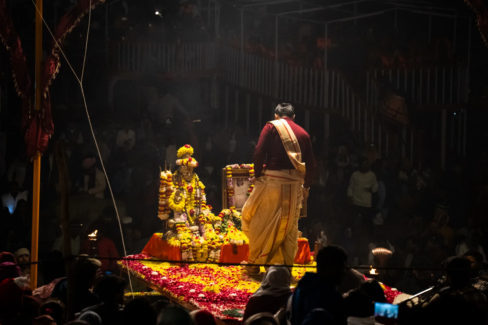 この写真は、インドのバラナシで撮影された宗教儀式の様子を捉えています。暗い背景に浮かび上がるように、鮮やかな色彩が印象的です。中央には、花で飾られた神像が置かれ、その周りには花びらが敷き詰められています。神像の横には額に入った写真が置かれています。神像の前に立つ男性は、伝統的な衣装を身に着けており、儀式を執り行っているようです。周囲には多くの人々が集まっており、儀式の様子を見守っています。写真全体は、宗教的な雰囲気と神秘的な空気に満ちています。煙のようなものが立ち込めており、幻想的な雰囲気を醸し出しています。