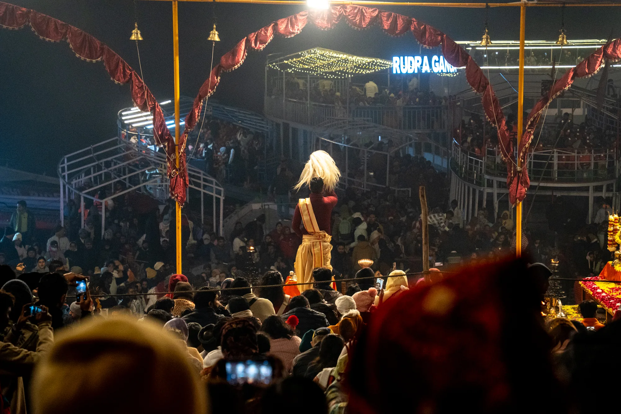 写真はインドのヴァラナシで撮影された、夜の風景を捉えています。手前には多くの人々が集まっており、その多くが後ろ姿で、頭や肩の一部が見えています。一部の人はスマートフォンで写真を撮っているようです。人々の中には、赤と金色の衣装を身につけ、頭に白い飾りをつけた男性が立っており、儀式のようなものを執り行っているように見えます。男性の背後には、人々が密集している高台があり、さらにその奥には「RUDRA GANGA」という文字がライトアップされた建物が見えます。建物の周りにも多くの人が集まっているようです。写真全体は暗いトーンで、祭りのような活気と神秘的な雰囲気が混ざり合っています。手前に設置されたアーチ状の装飾と、吊り下げられたベルが、さらに独特の雰囲気を醸し出しています。