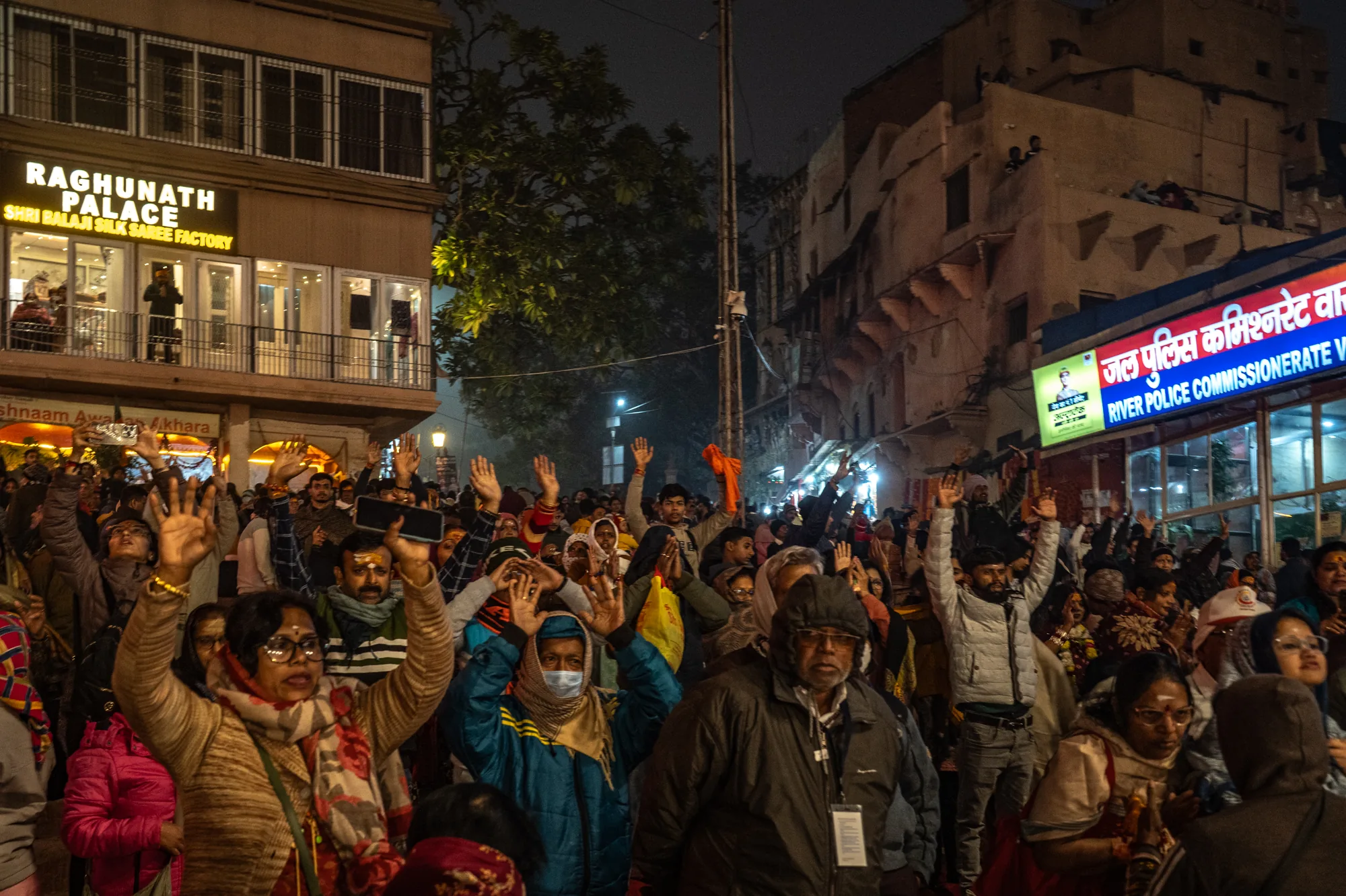 インドのバラナシで撮影された夜の写真です。多くの人々が集まり、手を高く上げています。何かの儀式か、宗教的な集まりに参加しているように見えます。前景には様々な服装をした人々が写っており、伝統的なインドの衣服を身に着けている人もいれば、現代的な服装の人もいます。数人はマスクを着用しています。  背景には、建物が並んでいます。「RAGHUNATH PALACE SHRI BALAJI SILK SAREE FACTORY」と書かれた看板が見えます。また、「जल पुलिस कमिश्नरेट वा(RIVER POLICE COMMISSIONERATE V)」と書かれた警察署のような建物も見えます。建物の窓からは明かりが漏れており、夜の暗さを照らしています。写真全体はやや暗く、群衆の熱気と宗教的な雰囲気を伝えています。