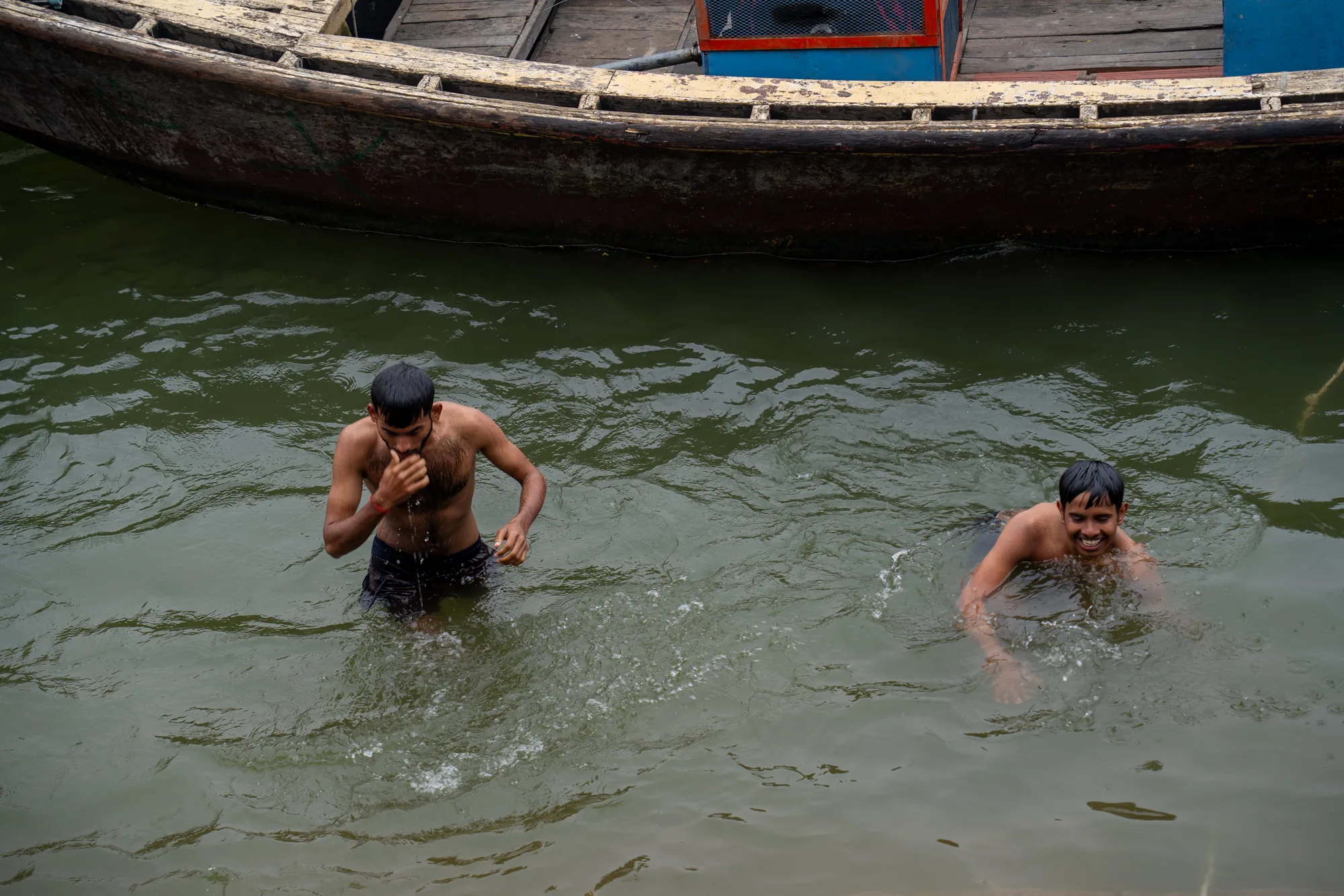 インドのバラナシで撮影された写真です。水面に2人の男性が浮かんでいます。一人は肩まで水に浸かり、もう一人は首から上だけが出ています。二人とも顔を上げ、カメラの方を見ています。水面には、彼らの動きによってできた波紋が広がっています。水の色は緑がかっており、濁っているように見えます。背景には、古い木造のボートの一部が写っており、歴史を感じさせる風合いです。ボートの縁は白く塗られていますが、剥がれかけており、長い年月を感じさせます。全体的に、この写真はバラナシの日常風景の一部を切り取ったものであり、聖なる川での沐浴の様子を捉えています。