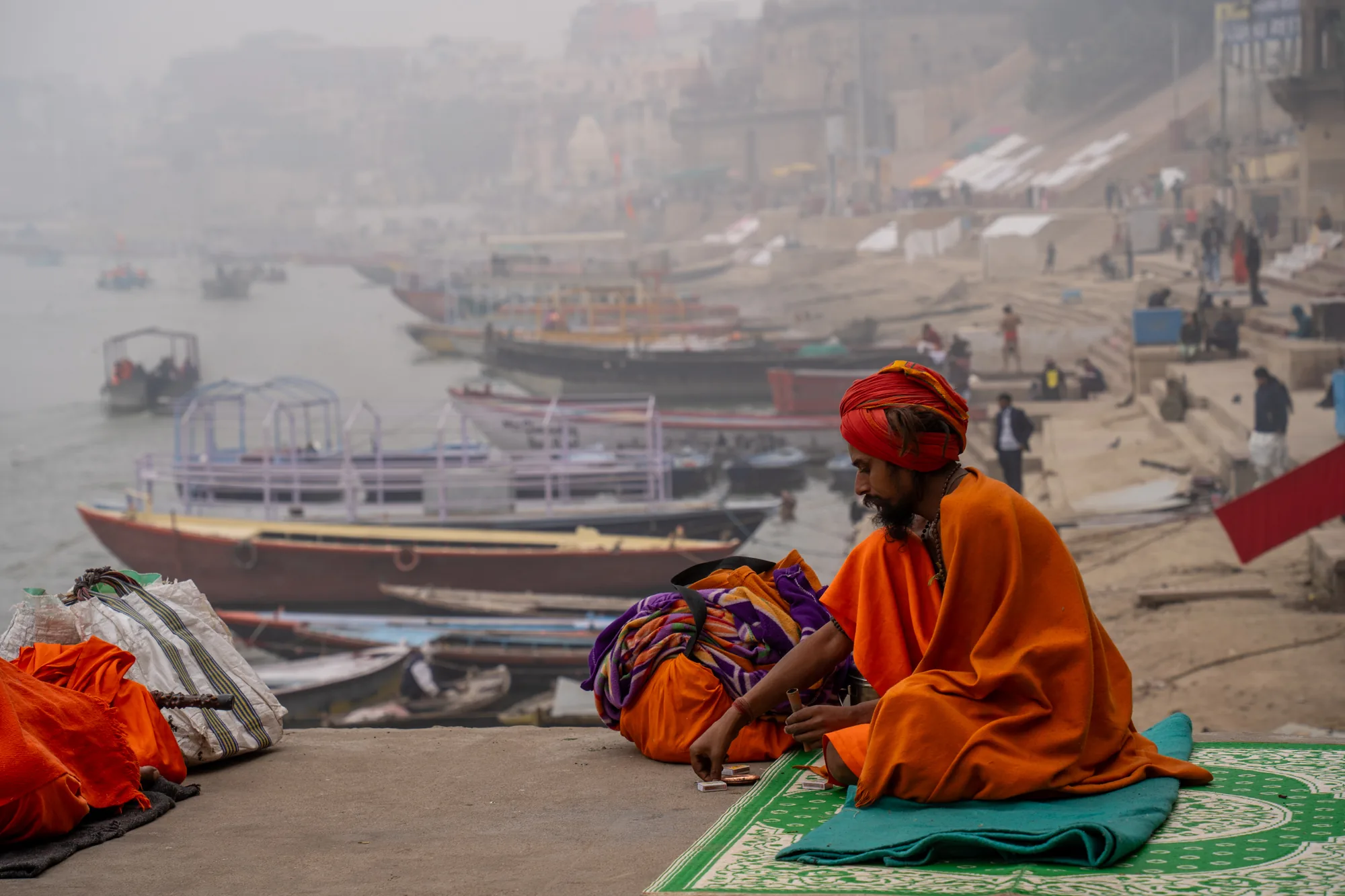 写真はインドのバラナシで撮影されたものです。手前には、オレンジ色の布をまとった男性がターバンを巻いて座っています。彼は緑色のマットの上に座り、何かを作っているようです。彼の横には、荷物や布などが置かれています。  背景には、霧が立ち込めるガンジス川が広がっています。川には多くのボートが浮かび、河岸には建物が立ち並び、人々が行き交っています。霧のせいで遠景はぼやけていますが、活気ある街の様子が伺えます。写真全体は、落ち着いた雰囲気で、バラナシの日常風景を切り取った一枚となっています。オレンジ色の服を着た男性が座っている場所は一段高くなっており、手前にグレーの壁が見えます。