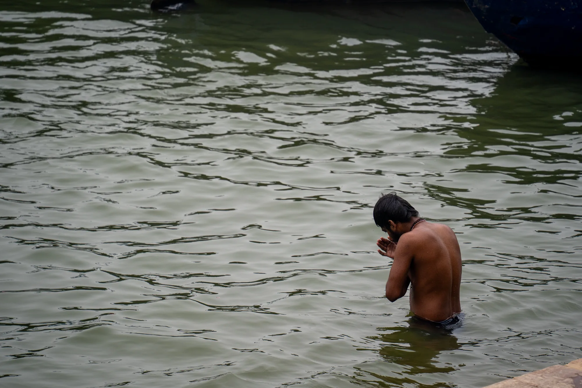 写真はインドのバラナシで撮影されたもので、ガンジス川と思われる水辺の様子が写っています。画面の大半は波紋が広がる水面で占められており、その水はやや濁った緑色をしています。水の表面は細かく波打ち、光を反射して複雑な模様を描いています。画面右側には、水に浸かった男性の後ろ姿が見えます。男性は上半身裸で、髪は短く、首にはネックレスのようなものを身につけています。男性は両手を合わせ、祈るような姿勢を取っており、敬虔な雰囲気が伝わってきます。画面右上に、船の一部と思われる濃い色の物体が写り込んでいます。全体的に静かで神聖な雰囲気が漂っており、バラナシの日常的な風景を捉えた一枚と言えるでしょう。