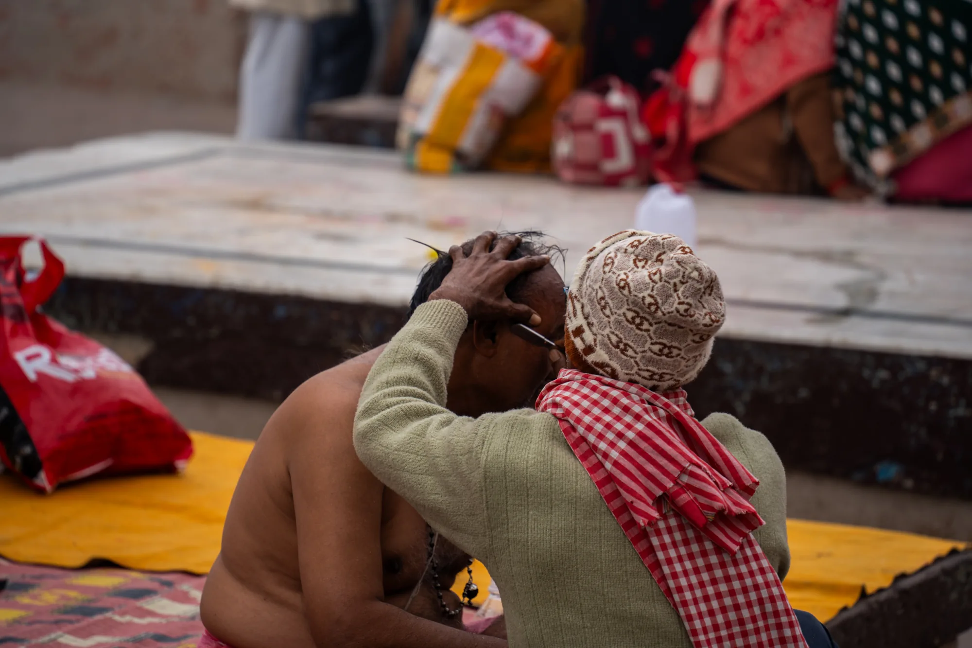 この写真はインドのバラナシで撮影されたものと思われます。写真には、上半身裸の男性が座っており、別の人がその男性の頭の毛を抜いている様子が写っています。毛を抜いている人は、ニット帽をかぶり、赤いチェックのストールを巻いています。男性の表情からは苦痛や抵抗は見られず、むしろ儀式の一部であるかのような静けさが感じられます。背景には、人々が座っている場所や荷物などが写り込んでおり、場所が神聖な場所であるか、もしくは何らかの儀式に関連する場所であることが推測できます。全体的に、この写真はバラナシの宗教的な側面や文化的な伝統を垣間見せるドキュメンタリー写真のような印象を与えます。