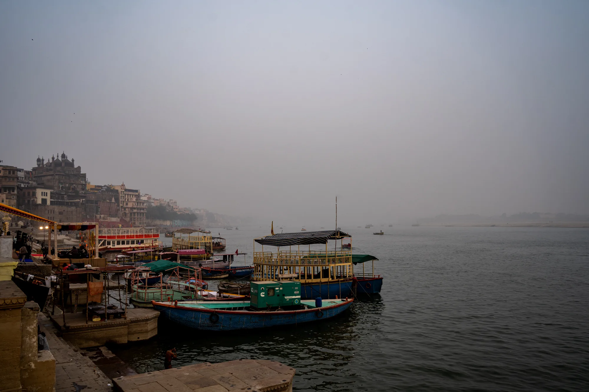 写真はインドのバラナシ（ベナレス）で撮影された、霧に包まれたガンジス川沿いの風景です。手前には石造りの岸壁があり、階段が水面に向かって続いています。水面には多くのカラフルなボートが停泊しており、赤、青、緑など様々な色が用いられています。一部のボートには屋根や装飾が施されており、観光客向けのボートのようです。岸壁には人々がおり、何らかの活動をしている様子が伺えます。  川の向こう側には建物が密集しており、歴史的な建築物が多く見られます。建物は霧によって霞んで見え、幻想的な雰囲気を醸し出しています。空は白く、全体的にどんよりとした天候であることがわかります。写真は落ち着いた色調でまとめられており、神聖な雰囲気と日常が混ざり合った、バラナシならではの風景を捉えています。全体的に静かで、どこか神秘的な雰囲気を持つ写真です。