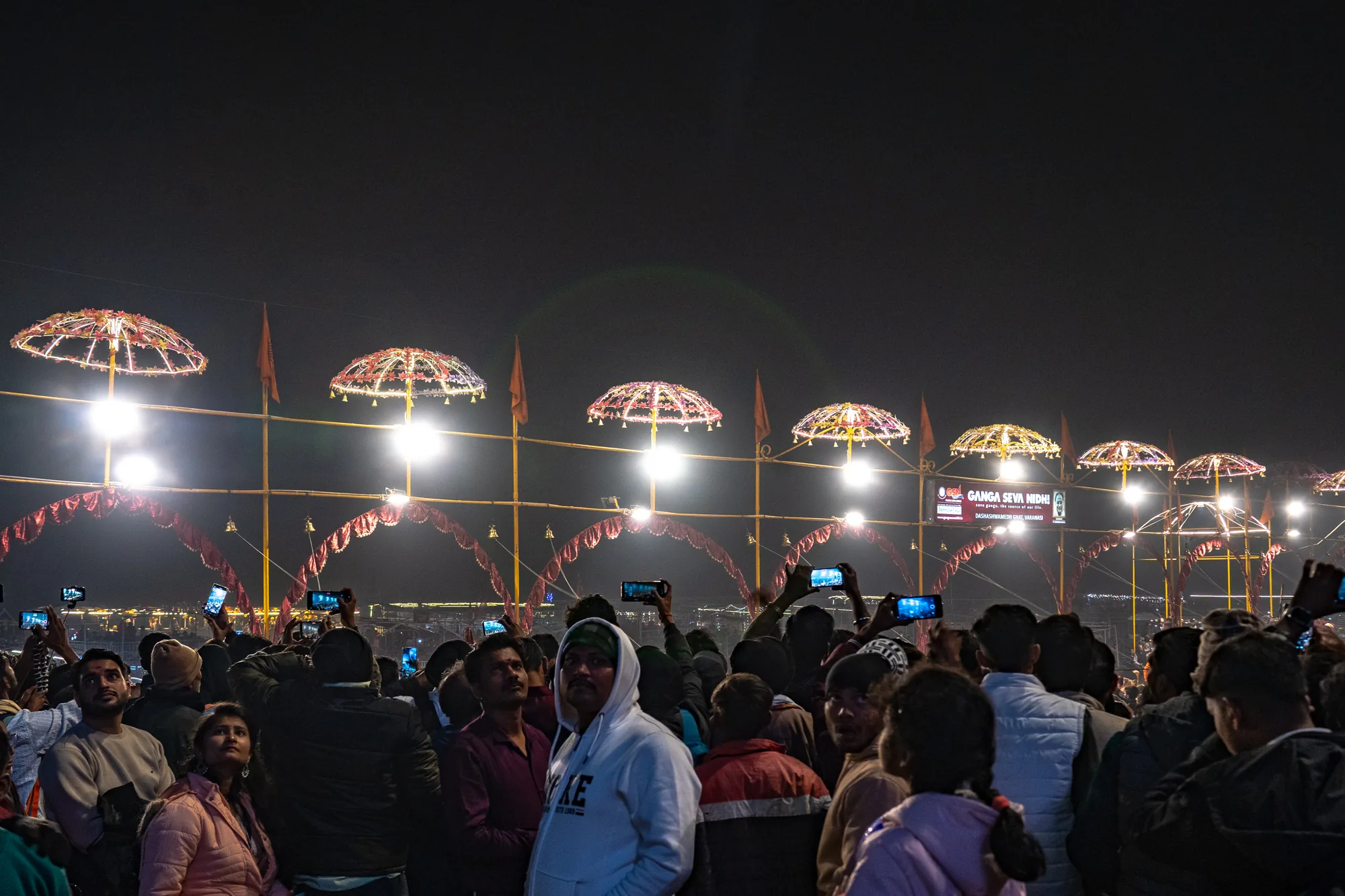 インドのヴァラナシで撮影された夜の写真です。暗闇の中、大勢の人々が何かを見上げています。多くの人がスマートフォンを高く掲げ、その光景を写真やビデオに収めようとしています。人々の視線の先には、ライトアップされた装飾的な傘が複数並んでおり、その下には赤いアーチ状の装飾も見えます。全体的に神秘的で、宗教的な雰囲気が感じられます。